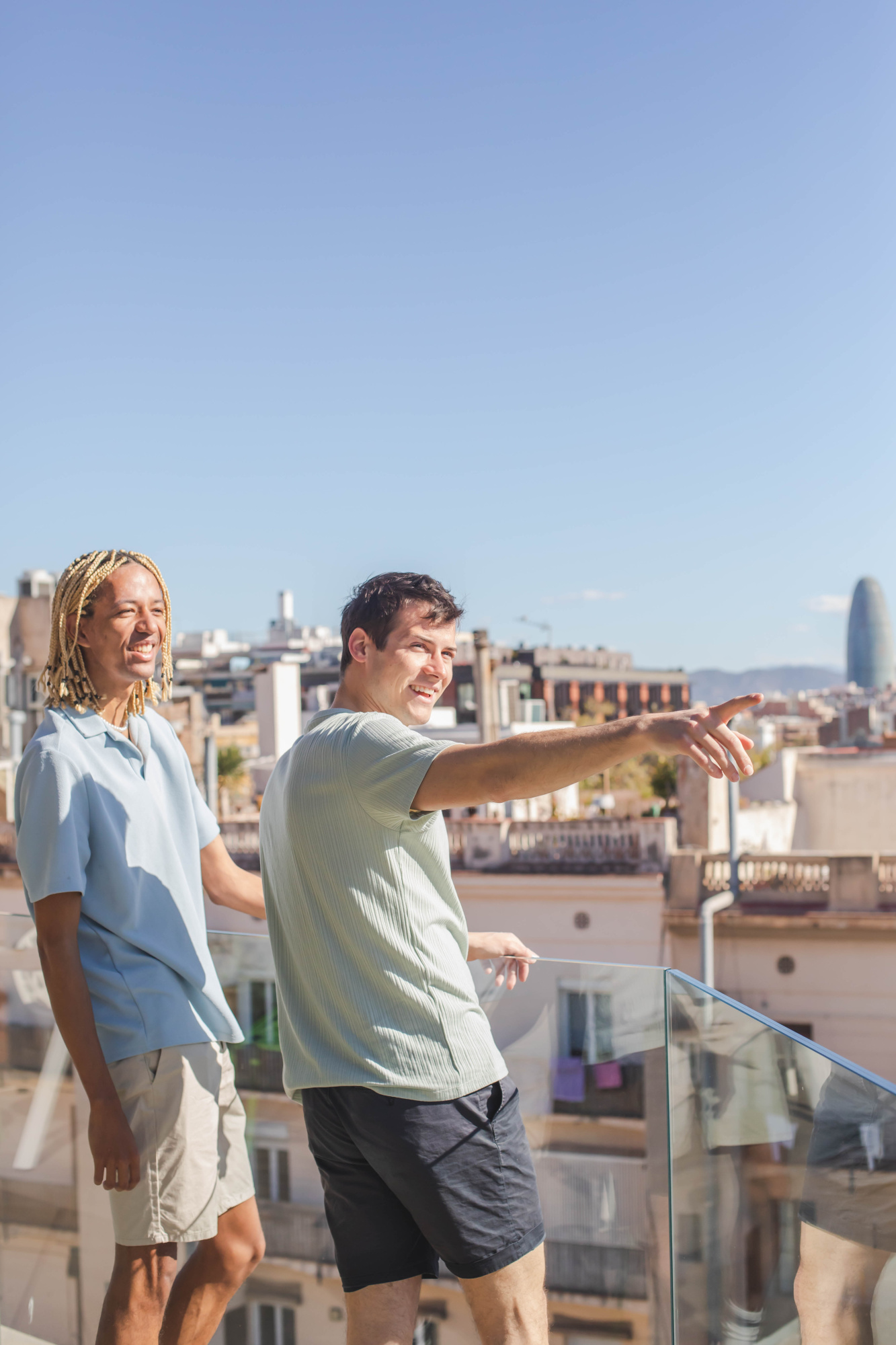 Dos personas caminan en una terraza con barandilla de vidrio y fondo urbano bajo el cielo despejado.