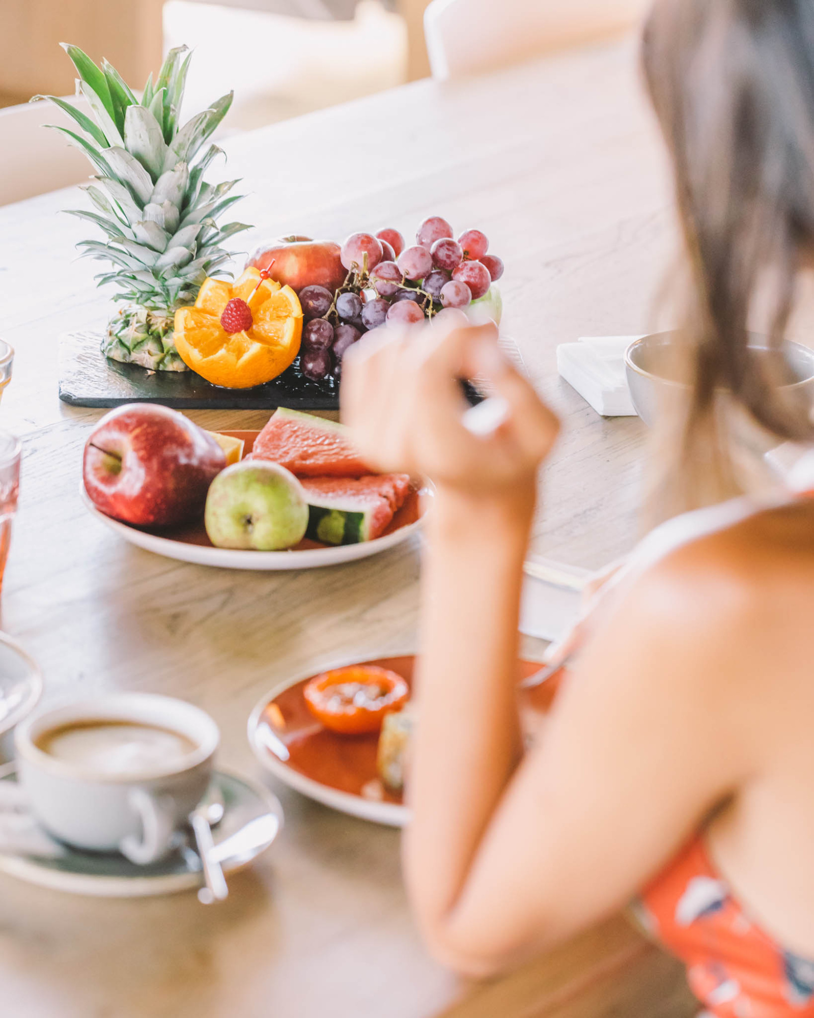 Una mujer desayuna junto a una mesa con frutas frescas y tazas de café.