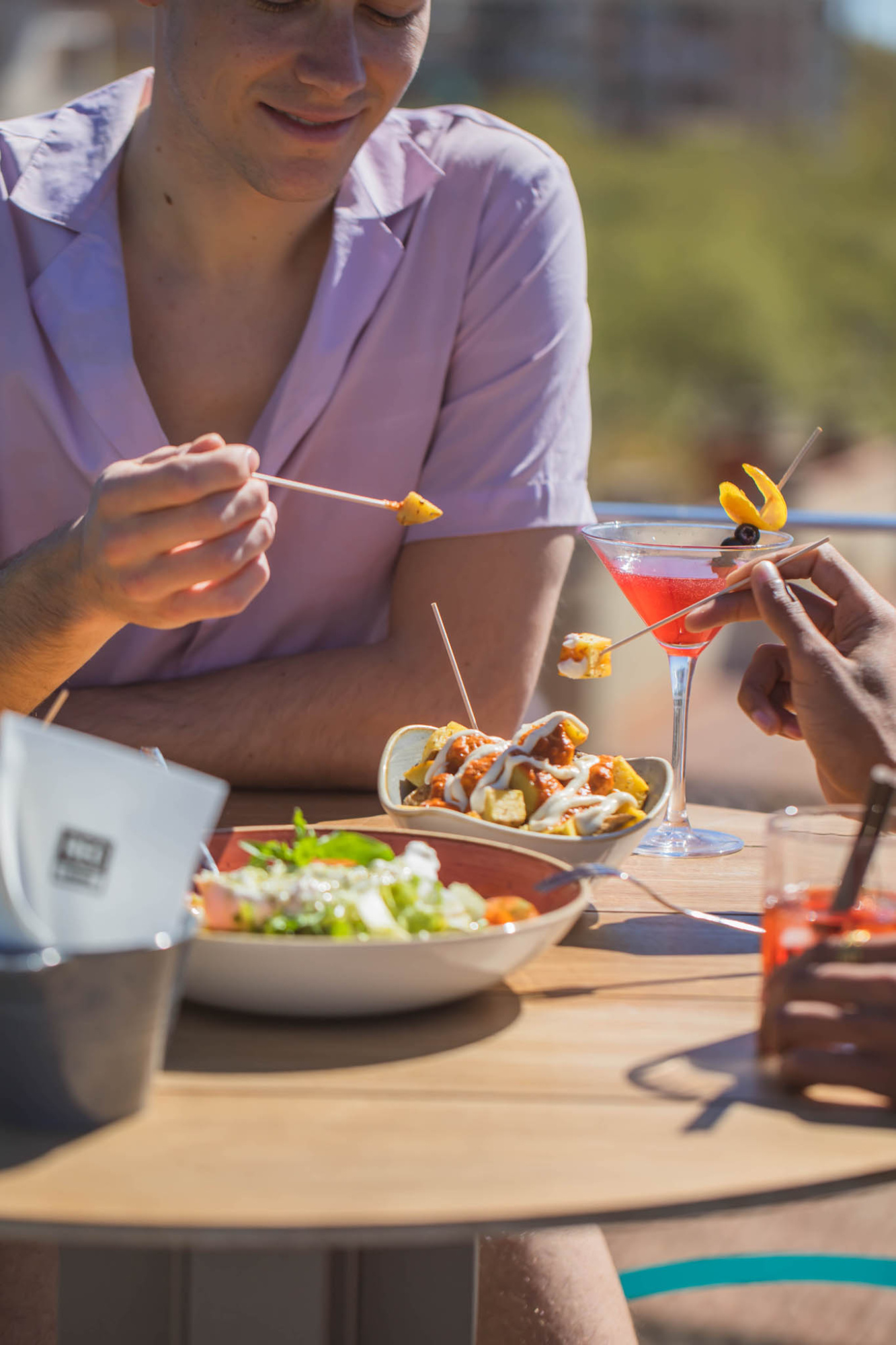 Varias personas comen al aire libre con platos y vasos sobre una mesa de madera.