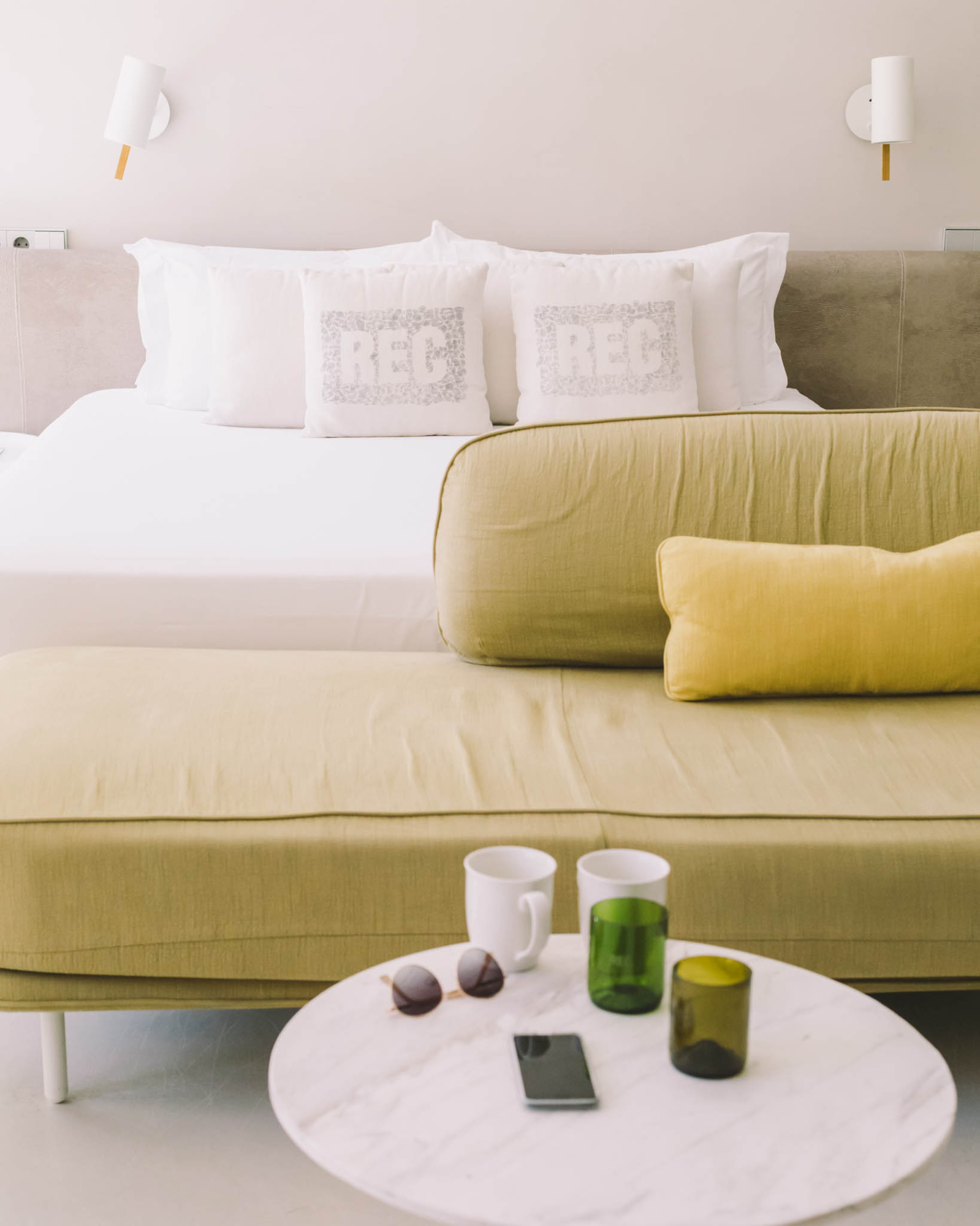 Se ve una cama con cojines blancos, una zona de estar y una mesa redonda con vasos y un teléfono.