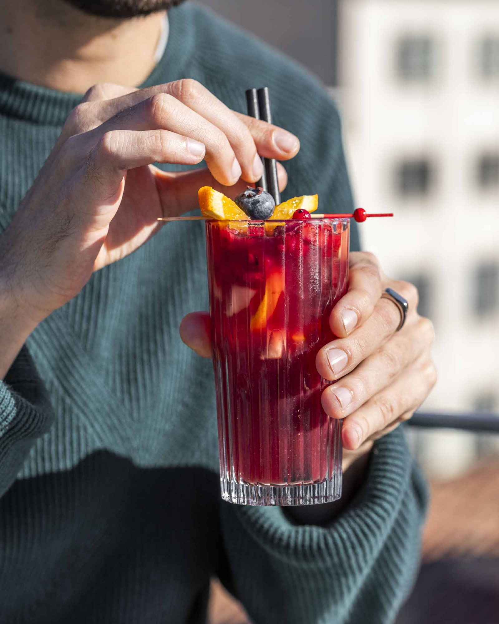 Sostiene un vaso alto con bebida roja y pajita mientras se ve un fondo desenfocado.