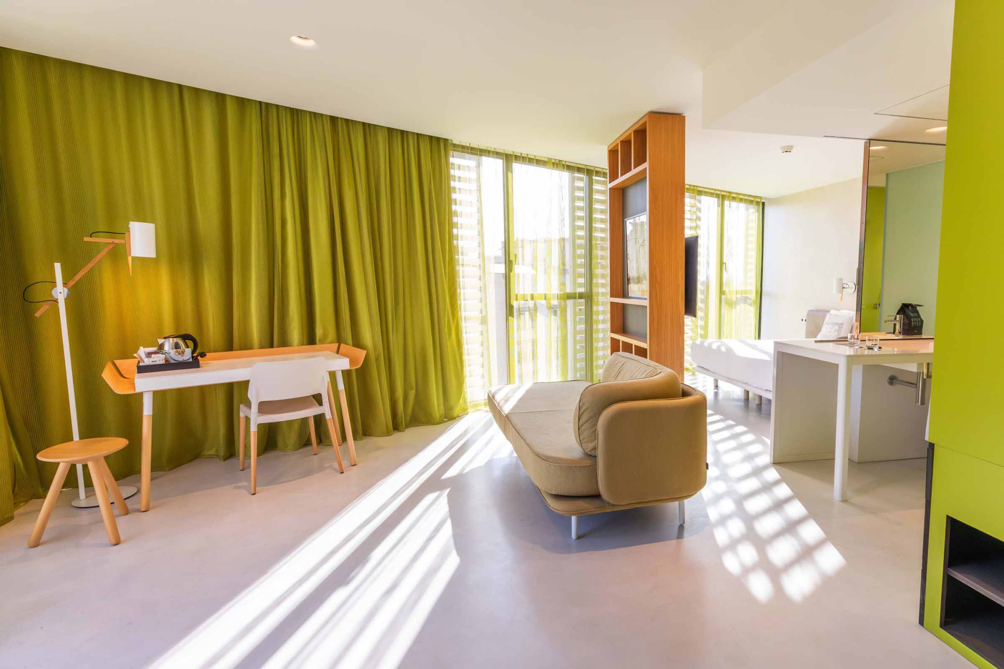 La imagen muestra una sala de estar con sillón, mesa, cortinas verdes y acceso a un balcón.