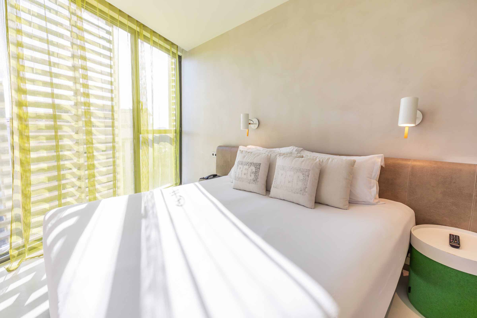 Se ve una cama con almohadas, una lámpara de mesa y una ventana con persianas junto a la pared.