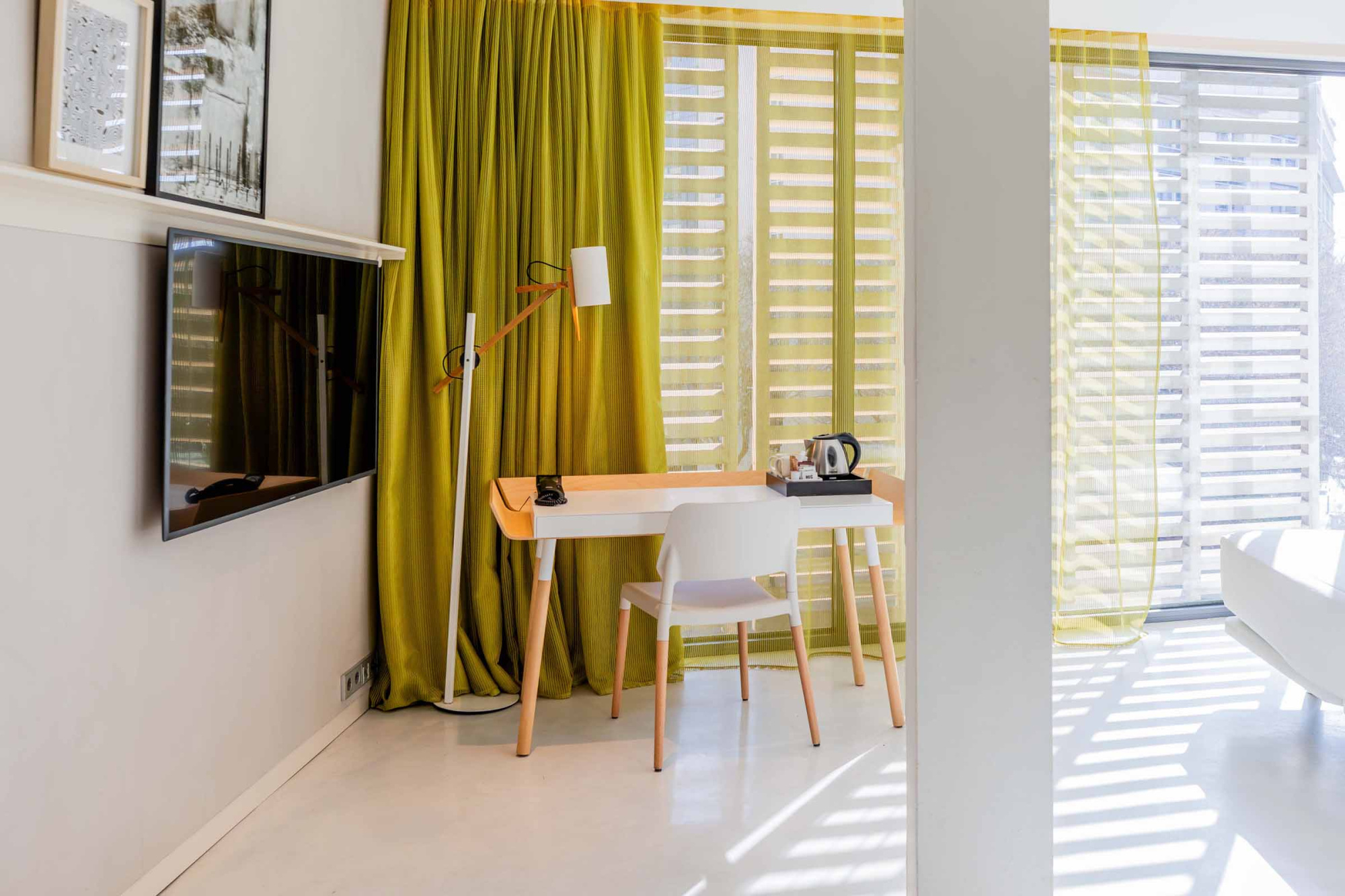 Se ve una mesa pequeña con dos sillas blancas junto a una ventana con cortinas verdes y persianas.