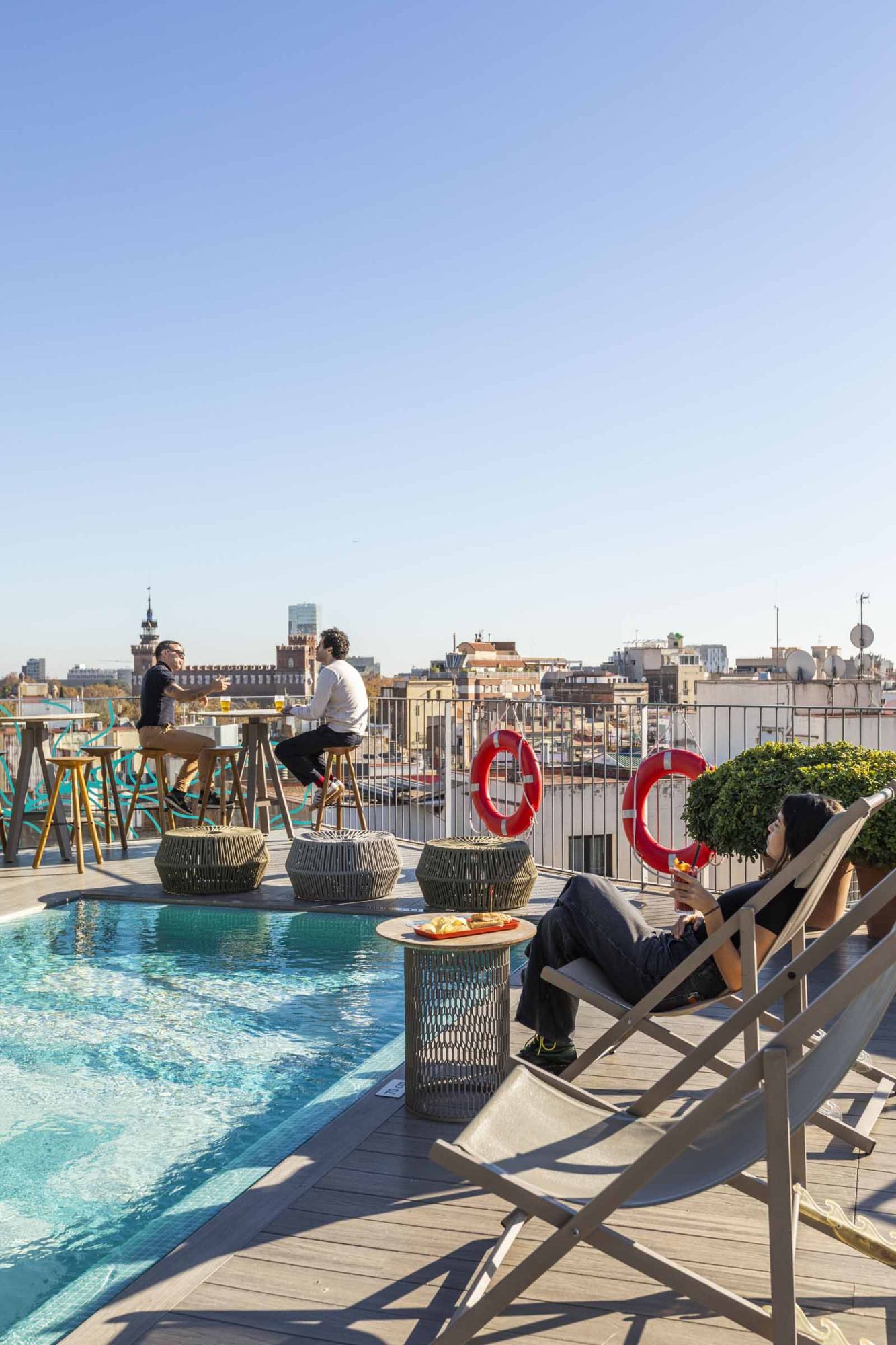 Se ve una terraza con piscina, tumbonas y sombrillas, con la ciudad al fondo.