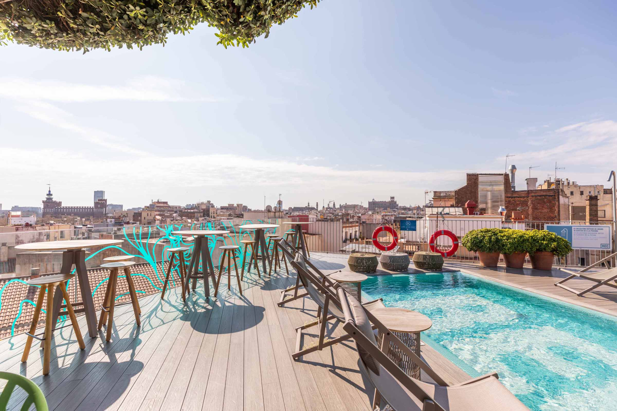 Se ve una piscina en una terraza con tumbonas, mesas y una vista de edificios al fondo.