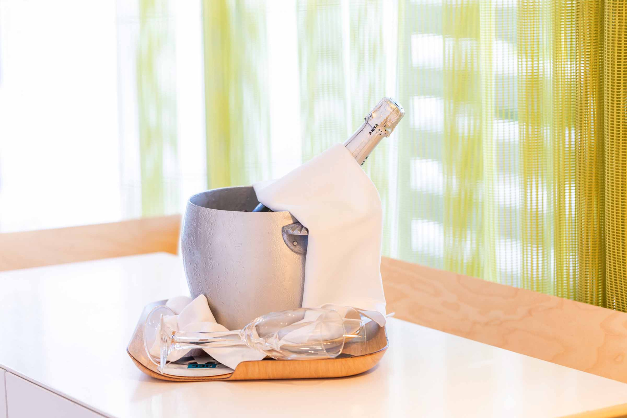 Se ve una mesa blanca con una botella y una copa sobre un mantel, junto a una cortina amarilla.