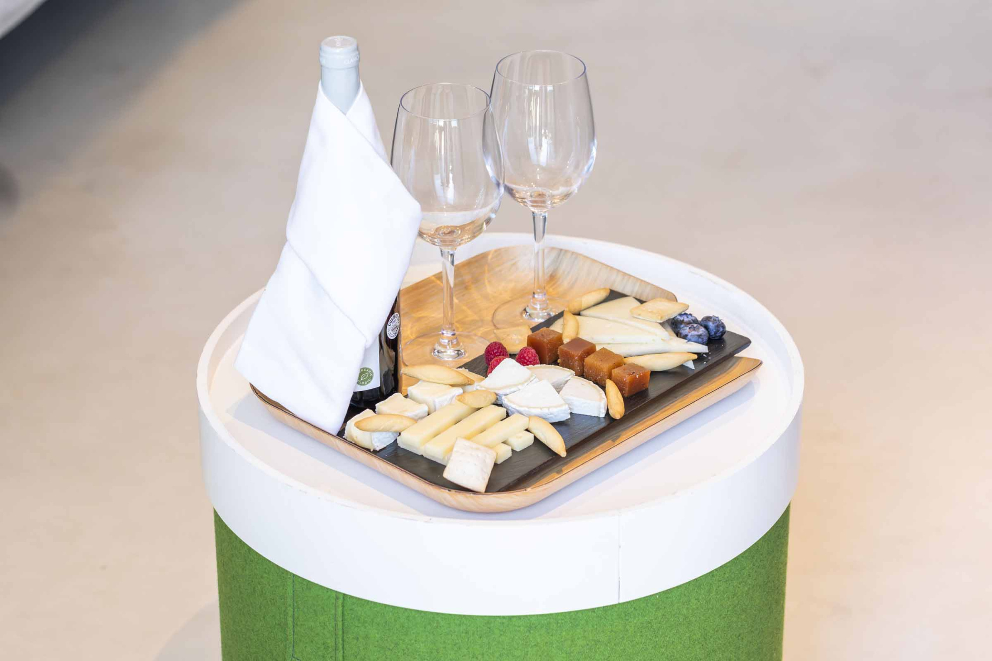 Se ve una mesa con una tabla de quesos, copas y una botella sobre un mantel blanco.