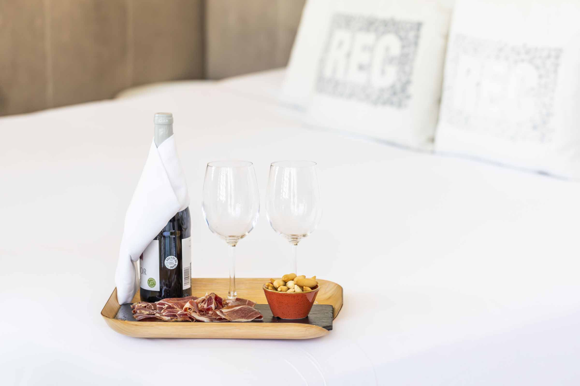 Se ve una mesa con una bandeja de madera, una botella, copas y aperitivos sobre un mantel blanco.