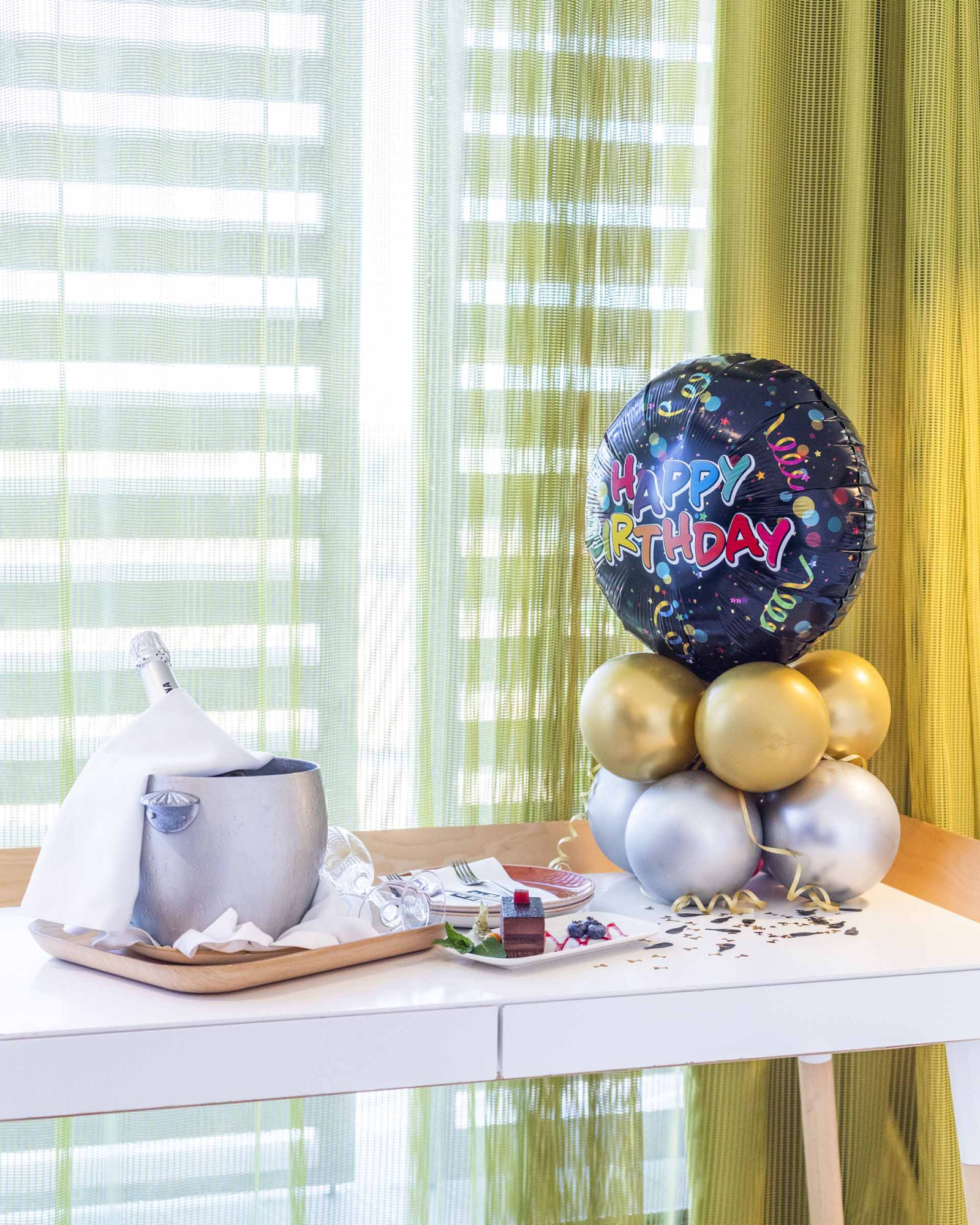 Se ve una mesa con globos y una decoración de cumpleaños junto a una ventana con cortinas.