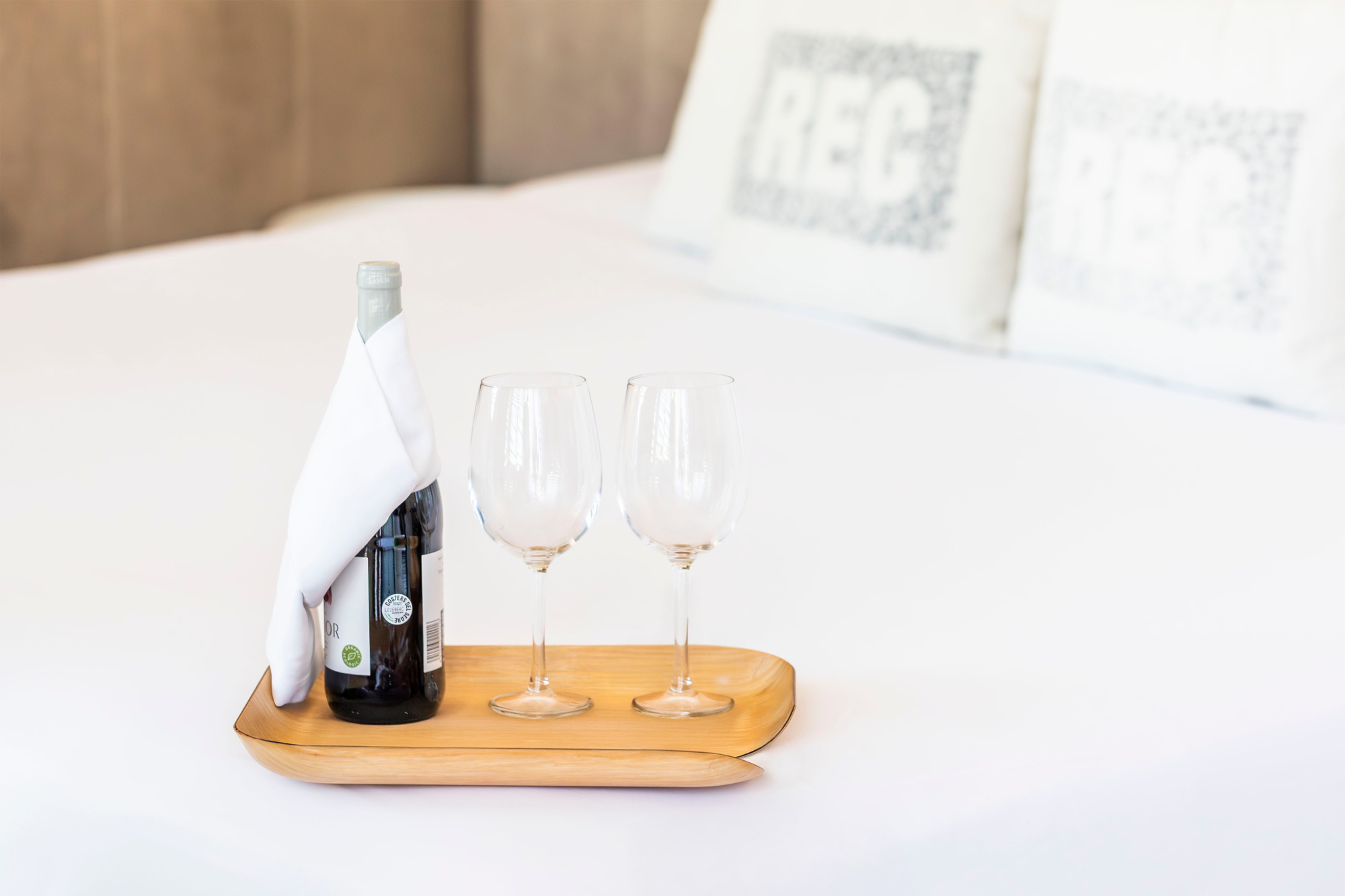 Se ve una mesa de madera con una botella y dos copas sobre una cama con cojines decorativos.