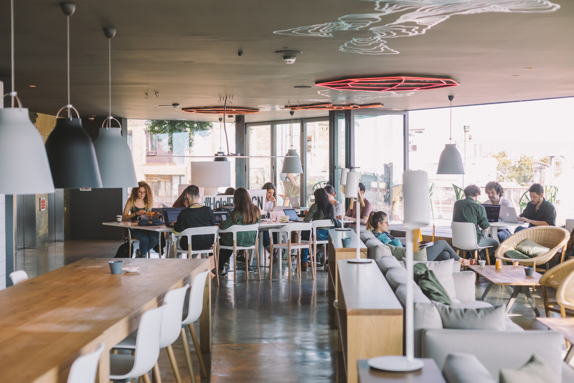 Se ve un comedor con mesas, sillas y personas sentadas junto a grandes ventanales.
