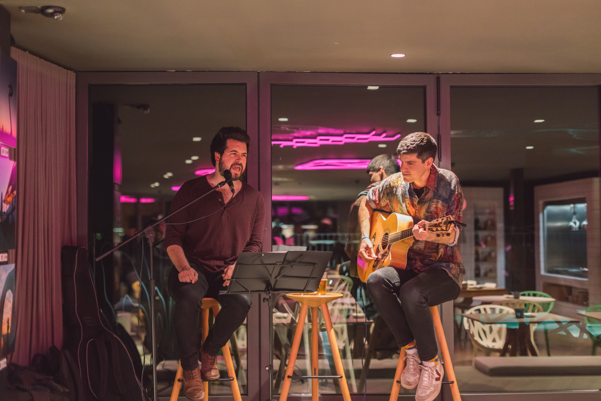 Dos músicos tocan instrumentos en un bar con luces de neón al fondo.