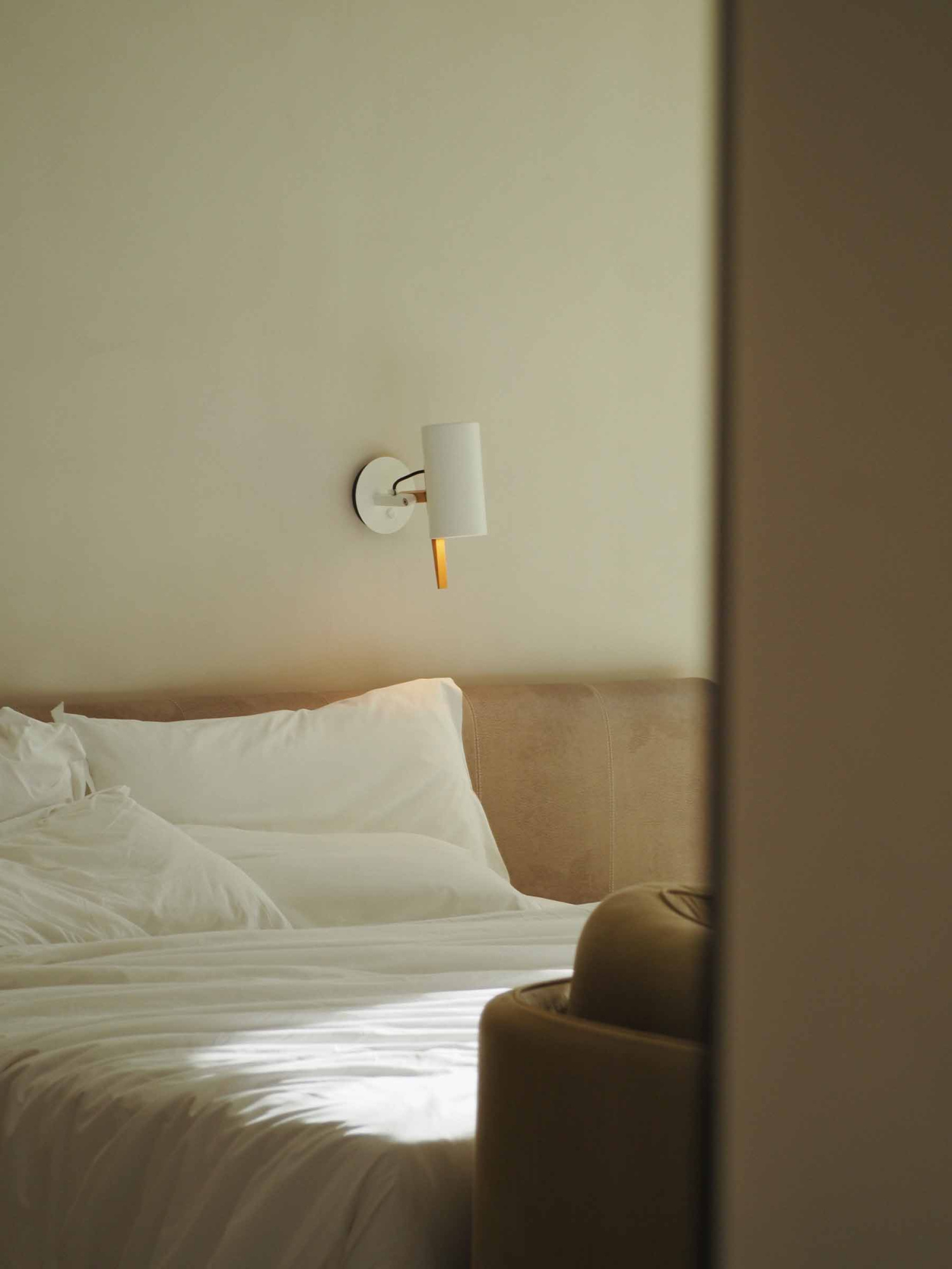 Se ve una cama con ropa de cama blanca y una lámpara de pared junto a la cabecera.