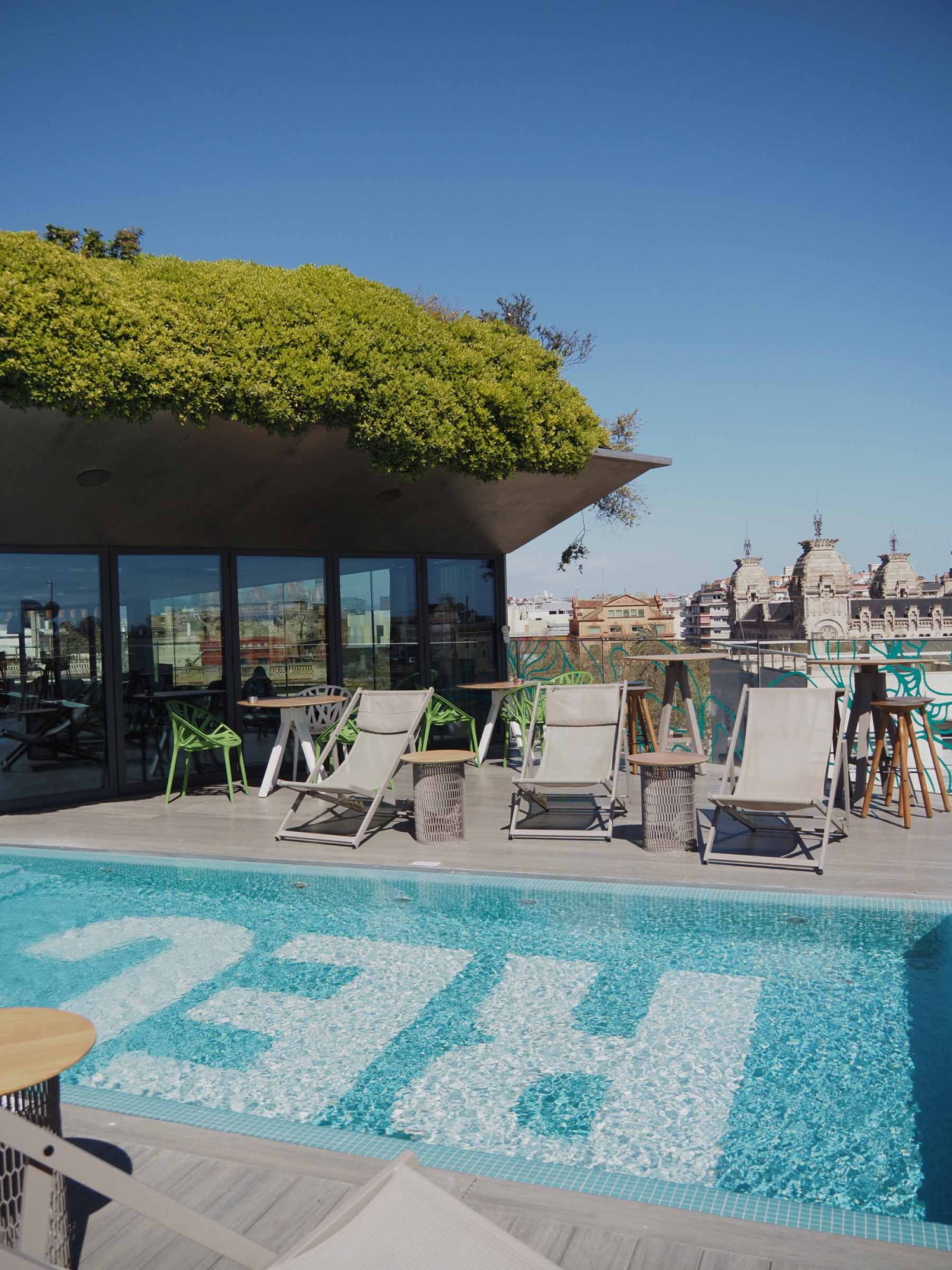 Se ve una piscina con tumbonas junto a una terraza con sillas y sombrillas bajo un toldo.