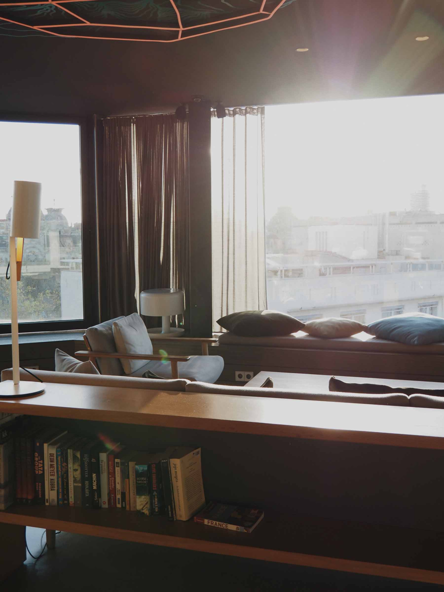 Se ve una cama con almohadas, cortinas oscuras y una lámpara junto a una repisa con libros.