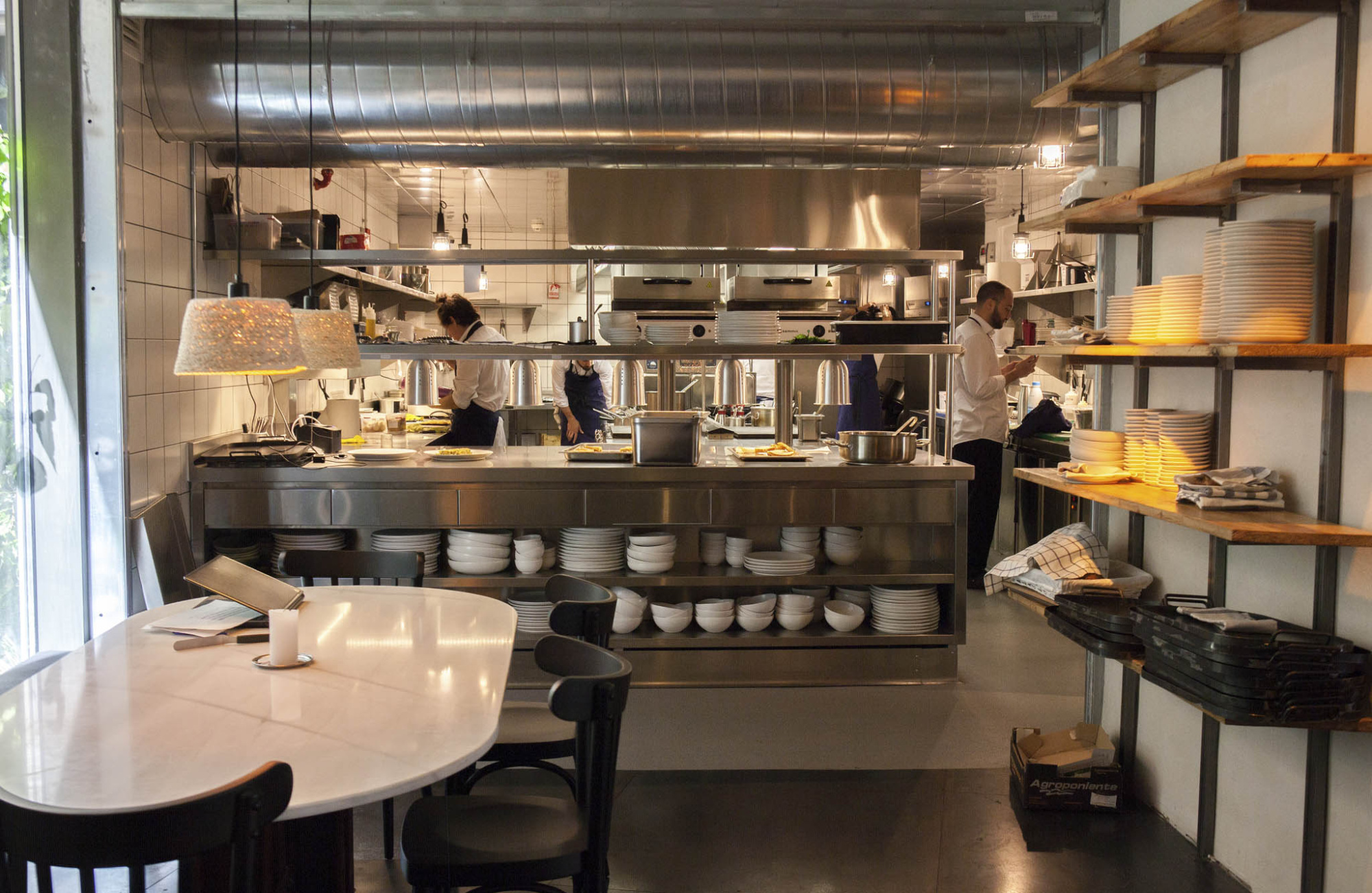 Se ve una cocina profesional con mesas de trabajo, utensilios y estanterías con platos.