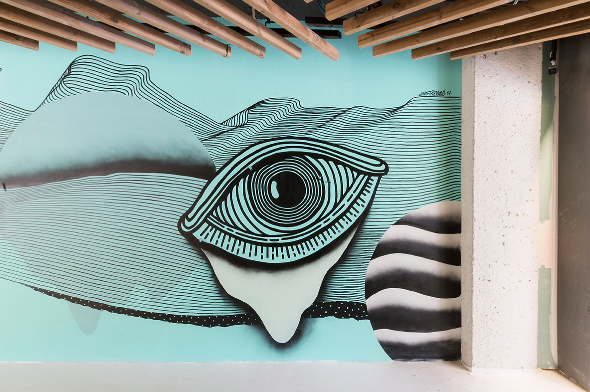 Muestra un mural con un ojo estilizado sobre una pared y una columna blanca al lado.