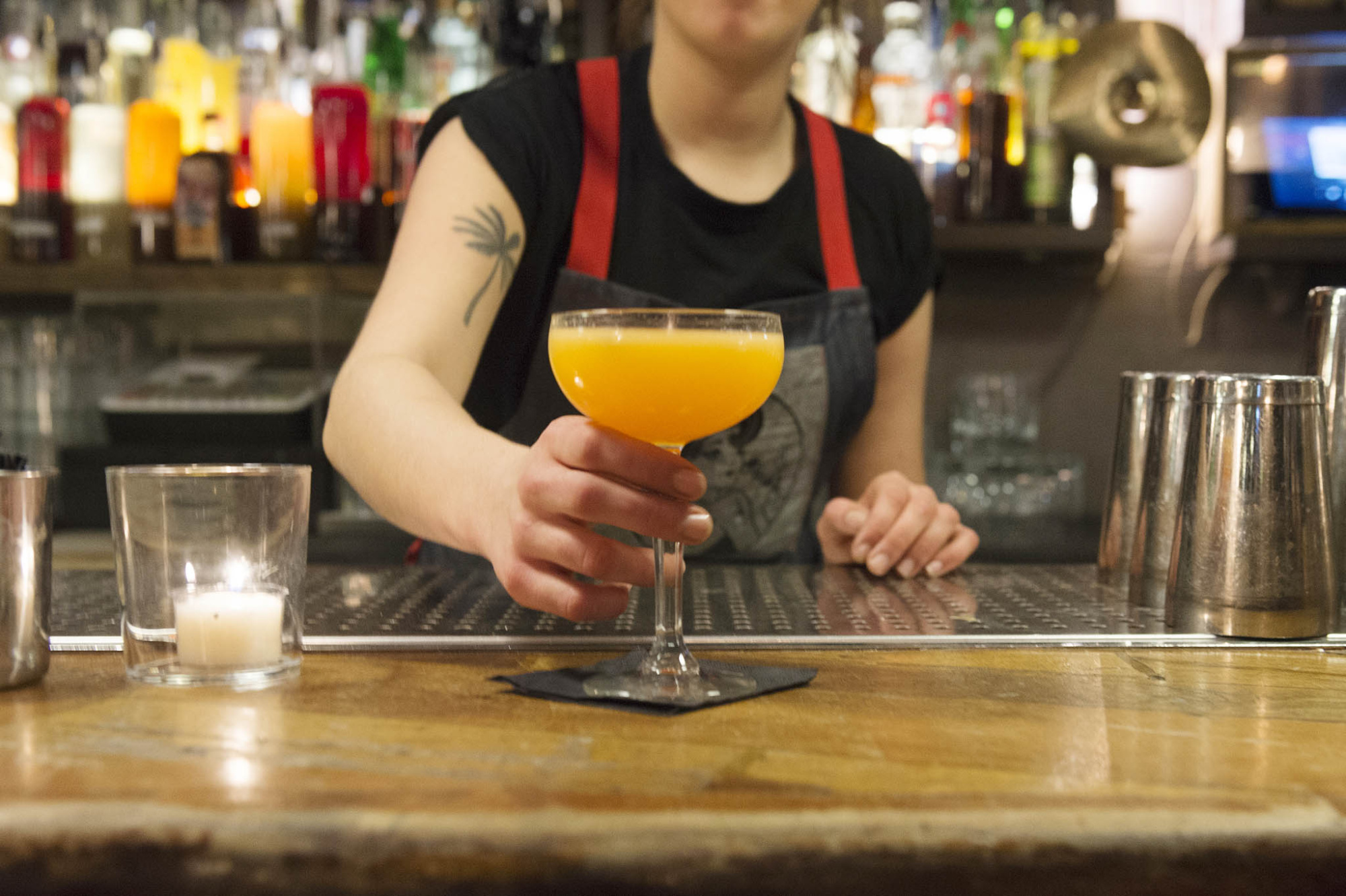 Una persona sostiene una copa con una bebida naranja en una barra de bar con botellas al fondo.