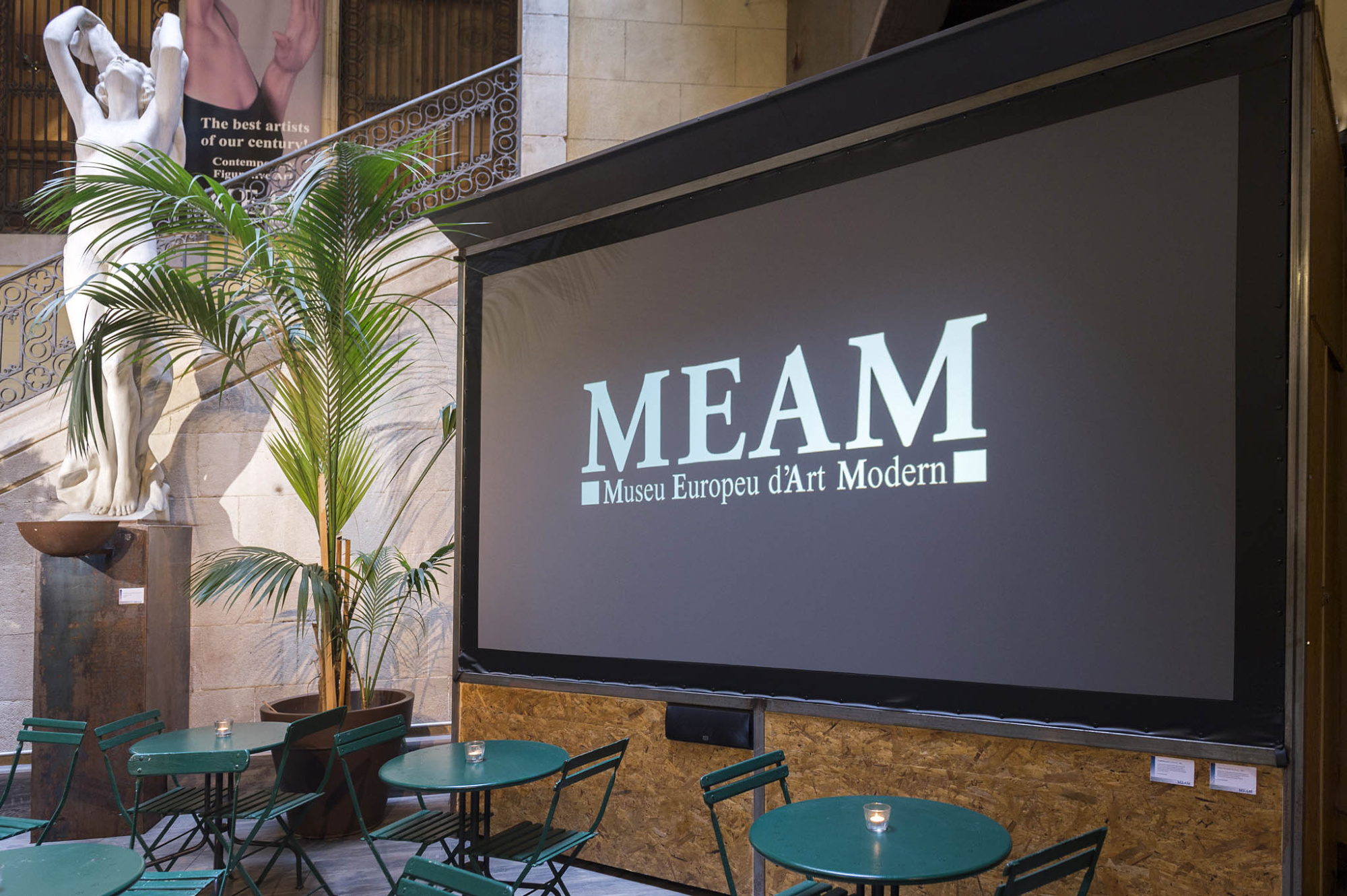 Se ve una pantalla con el texto “MEAM” y una zona exterior con mesas y sillas junto a una pared de ladrillo.