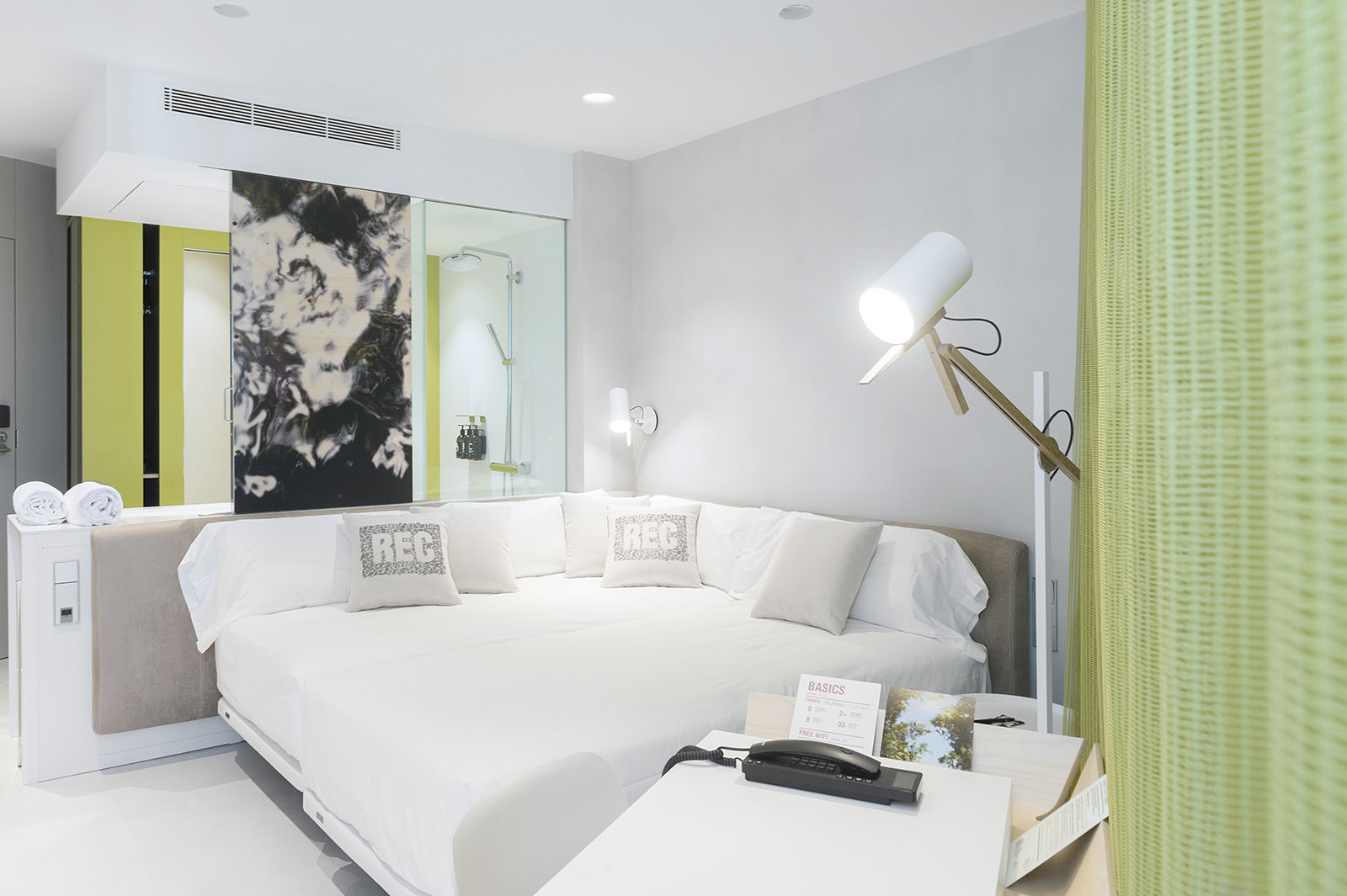 Se ve una cama doble con cojines, una lámpara de pie y un escritorio con bandeja sobre una habitación moderna.