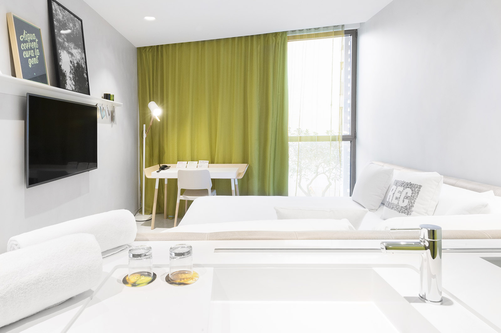 Se ve una cama doble, un escritorio con silla, una ventana con cortinas verdes y un televisor en la pared.