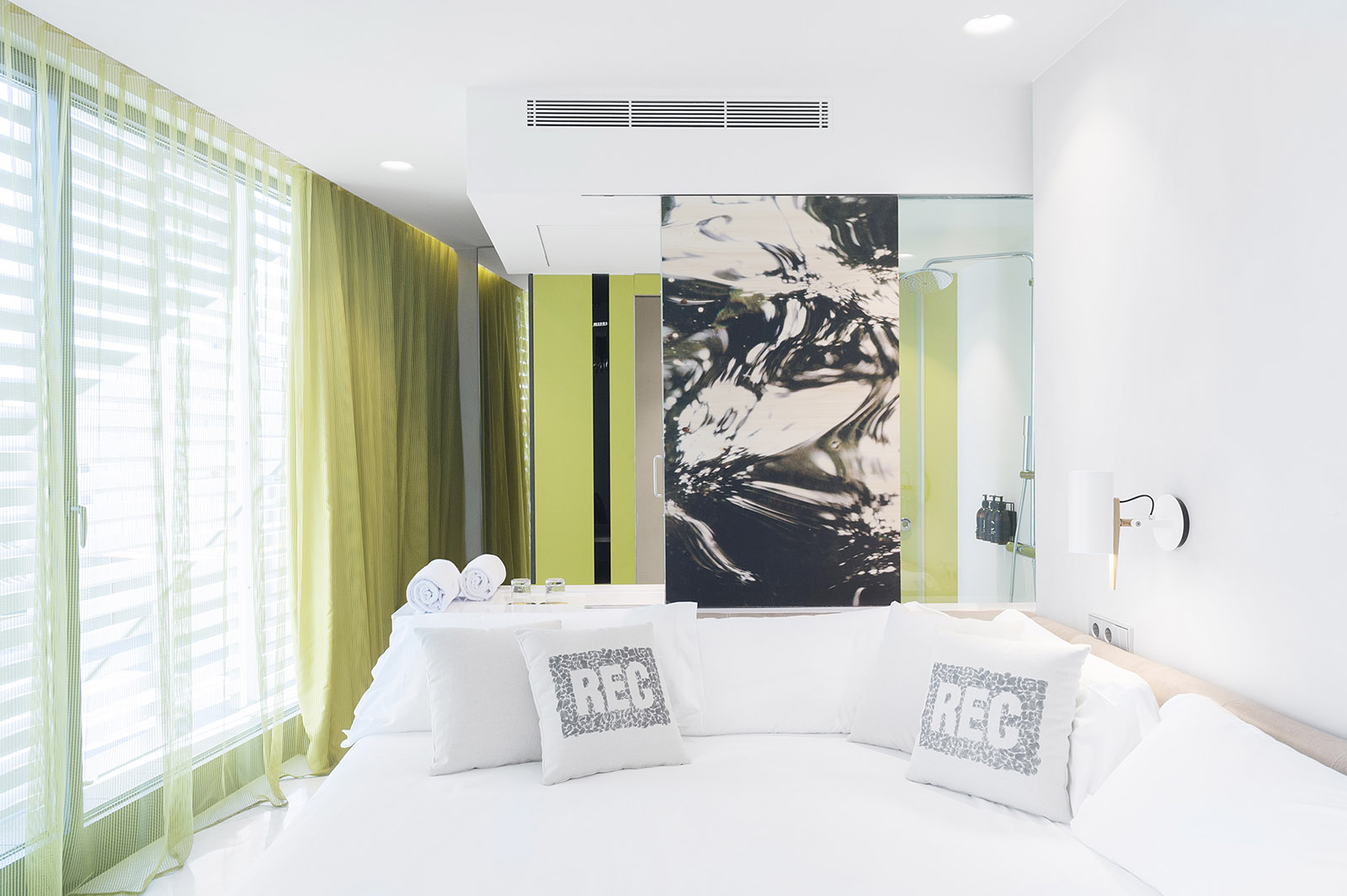 Se ve una cama doble con cojines y cortinas verdes junto a una pared con un cuadro grande.