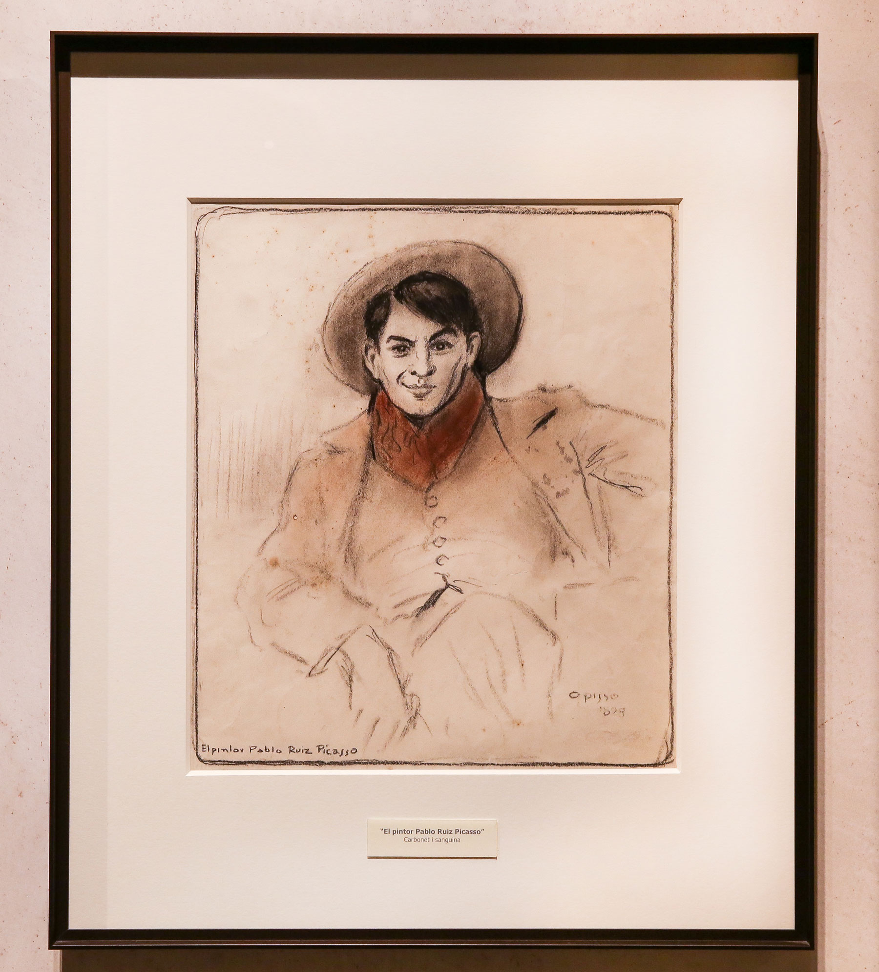 Se ve un cuadro enmarcado con un retrato de Pablo Picasso dibujado por Ricard Opisso y un fondo claro en una pared interior.