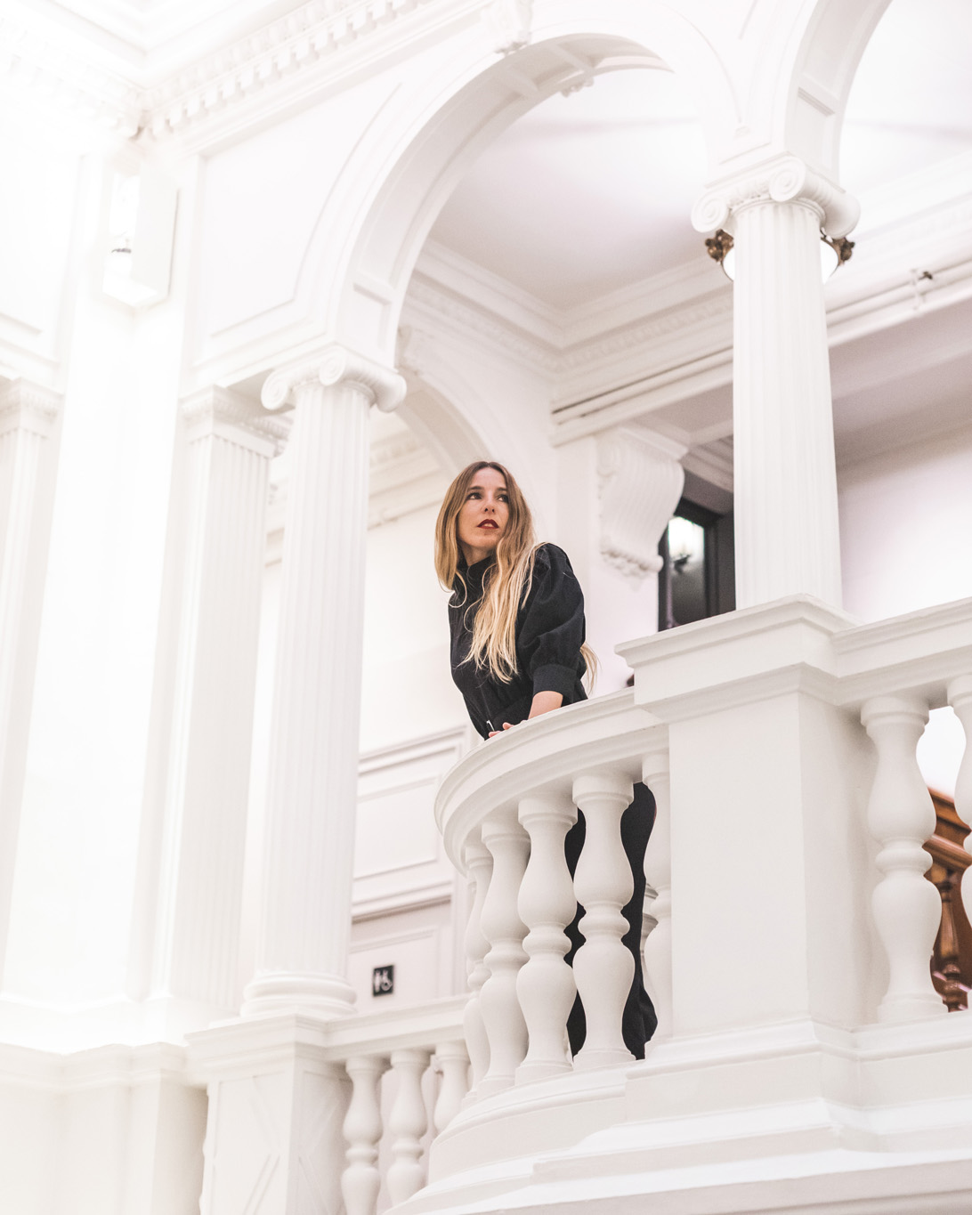 A woman poses on an interior balcony with a white railing and decorative columns.