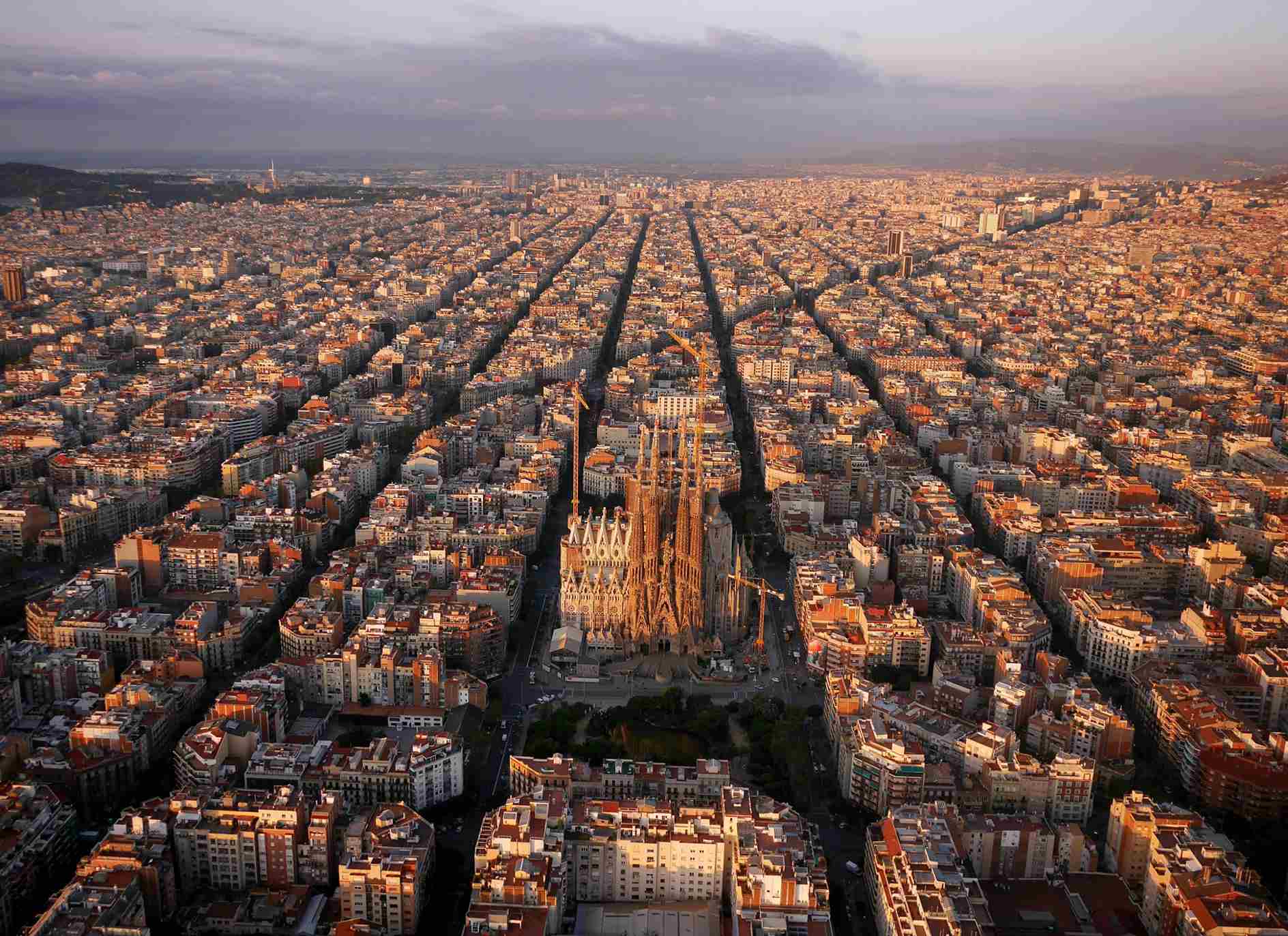 A cityscape of the Eixample is shown, with long streets and dense buildings beneath a cloudy sky.