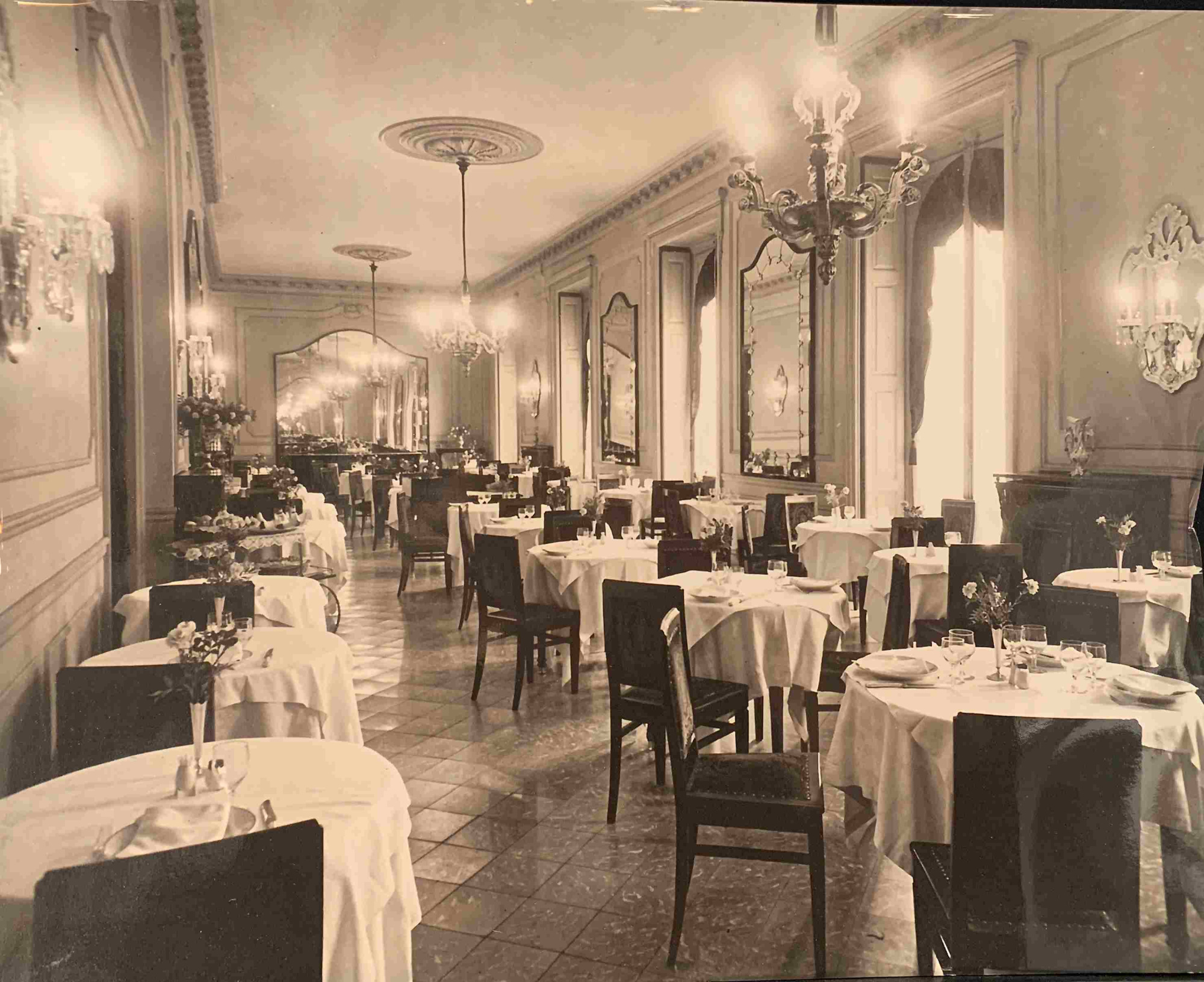 Se ve un comedor con mesas cubiertas y sillas alrededor, con lámparas colgantes y paredes decoradas.