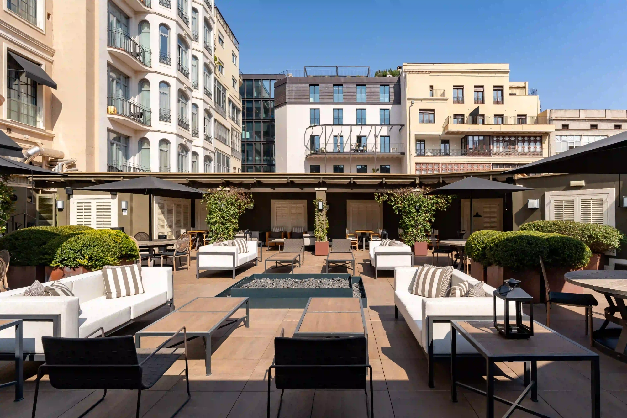Se ve una terraza exterior con mesas, sillas y tumbonas alrededor de una zona ajardinada.