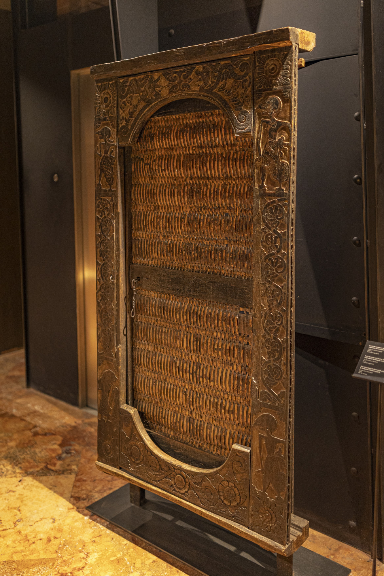Se ve un panel decorativo de mimbre con marco dorado sobre una base oscura en una sala interior.