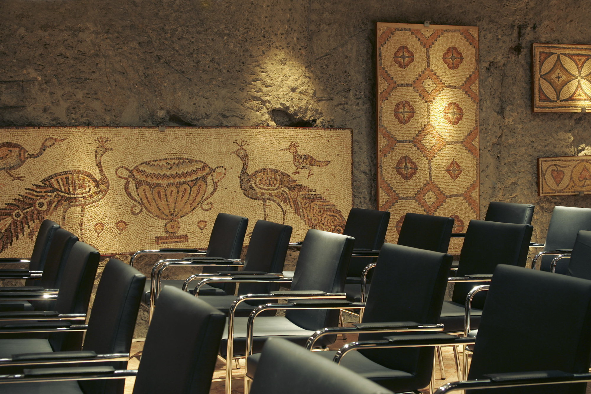Se ve una sala de reuniones con filas de sillas negras y mesas metálicas, con decoración de auténticos mosaicos romanos en las paredes.