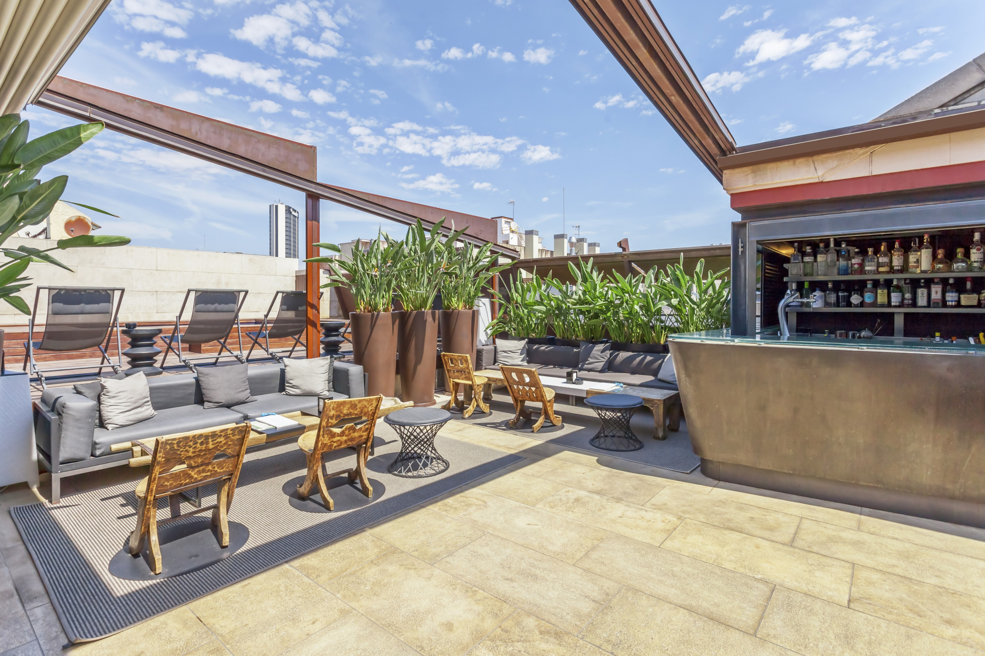 Se ve una terraza en la azotea con mesas y sillas, plantas y una barra con botellas.