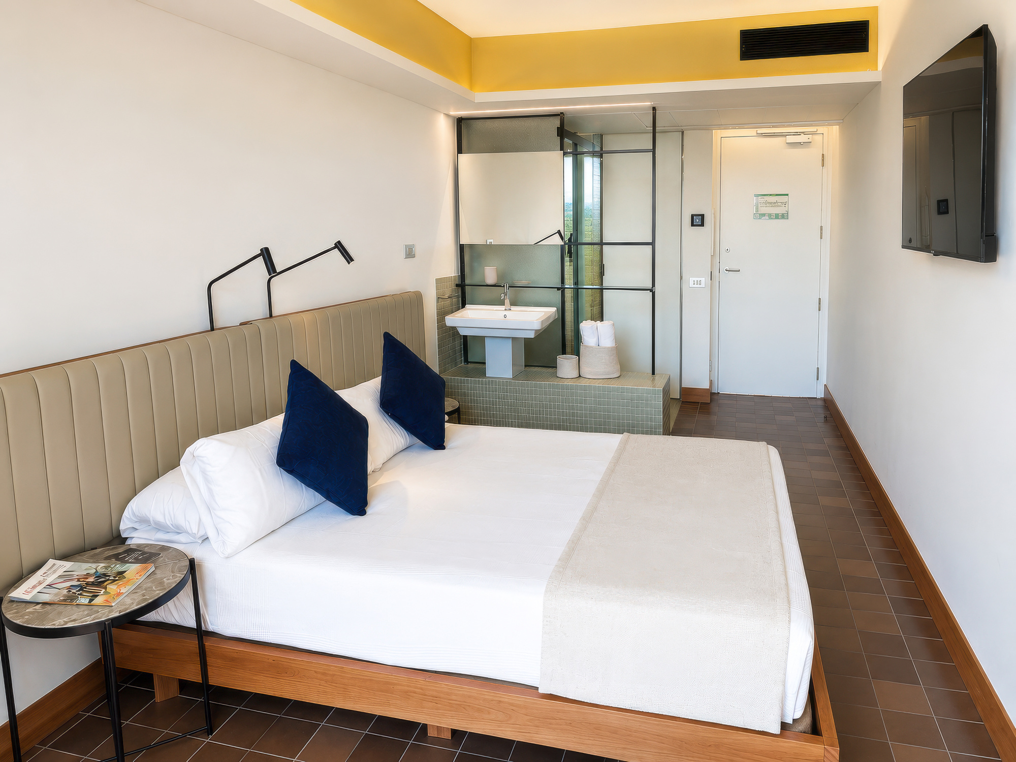 La imagen muestra una cama doble con almohadas azules y una lámpara, junto a un área con lavabo y acceso a una puerta.