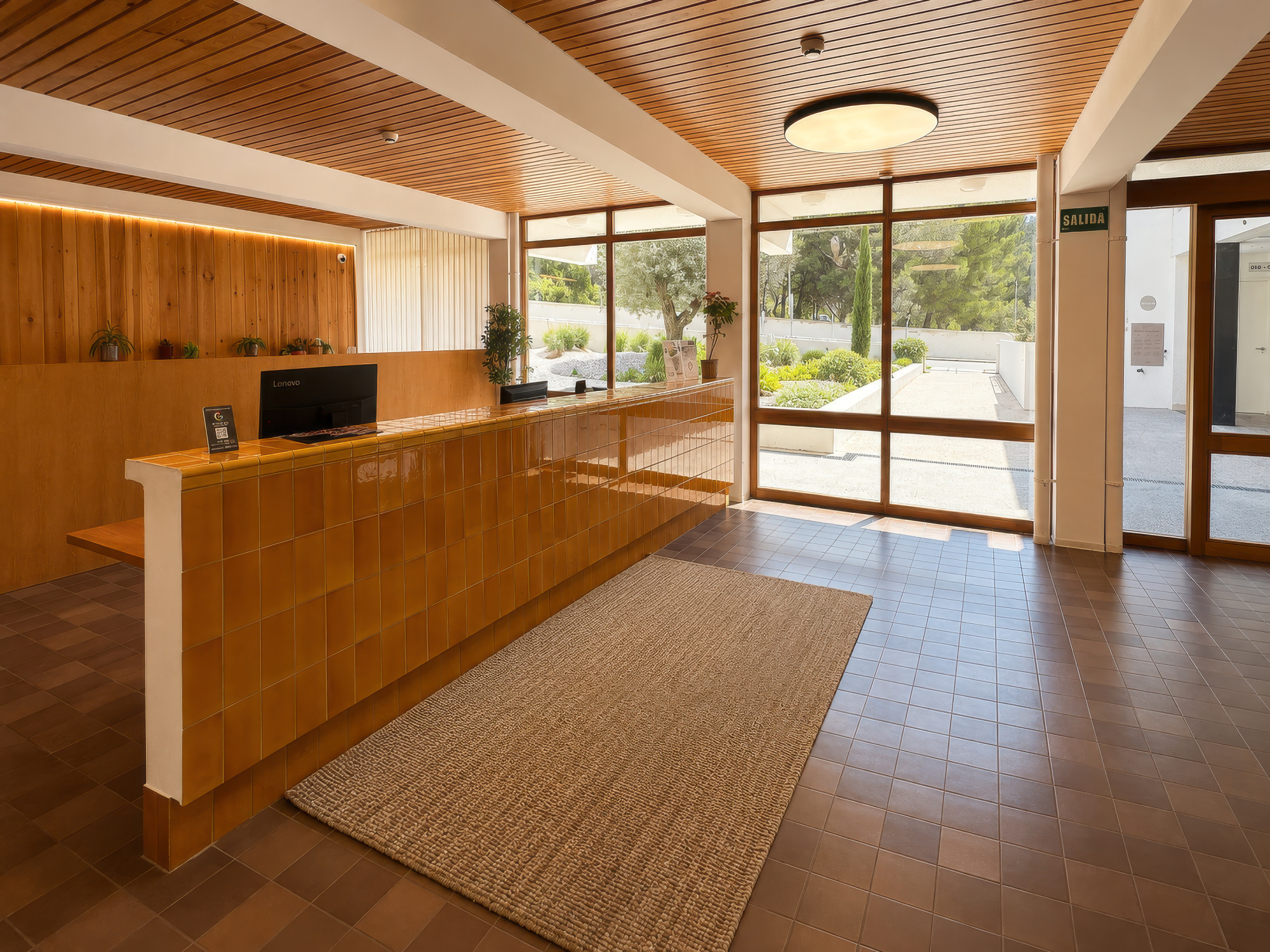 Se observa una recepción con mostrador de madera, una zona de estar y puertas de acceso con luz natural.