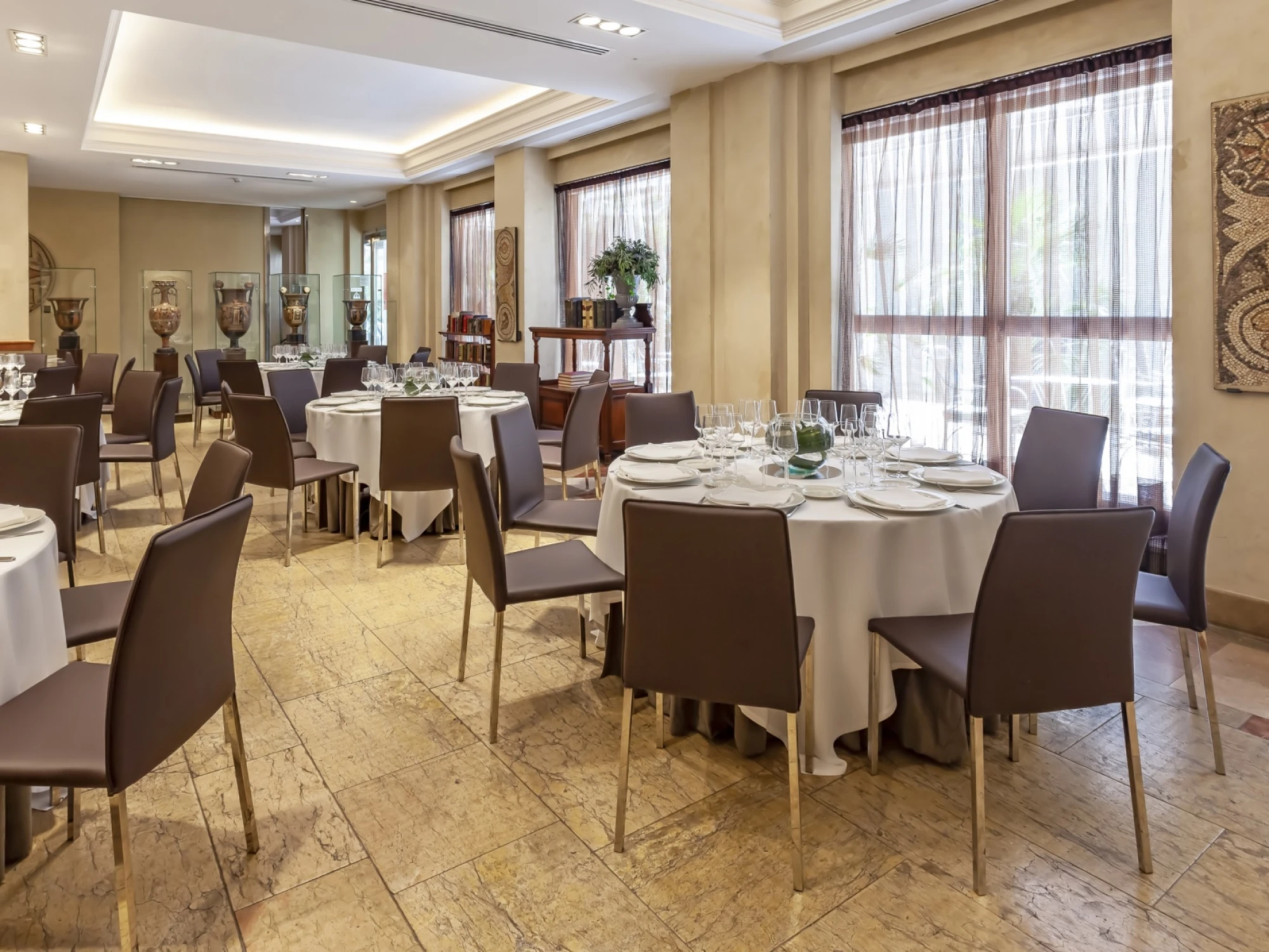 Comedor con mesas cubiertas con manteles blancos y sillas marrones junto a grandes ventanales.