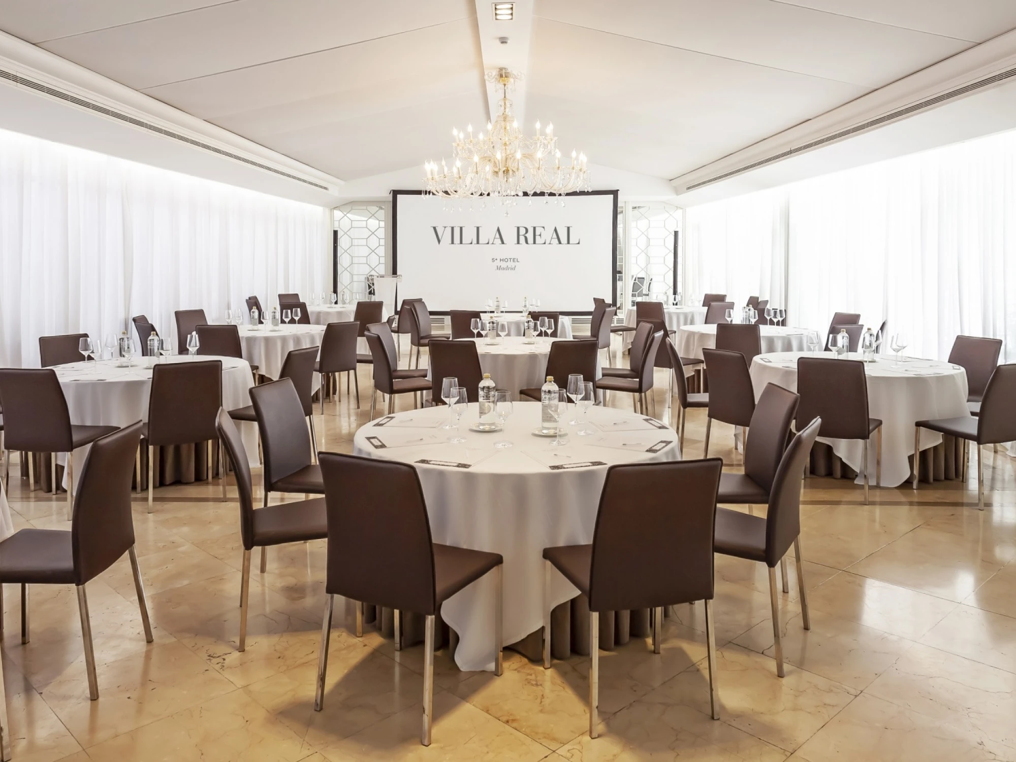 Se ve una sala de comedor con mesas redondas cubiertas con manteles blancos y sillas marrones.