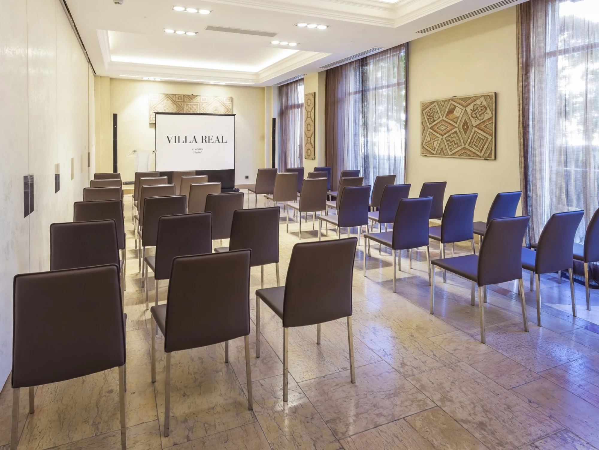 Se ve una sala de reuniones con filas de sillas y ventanas con cortinas.