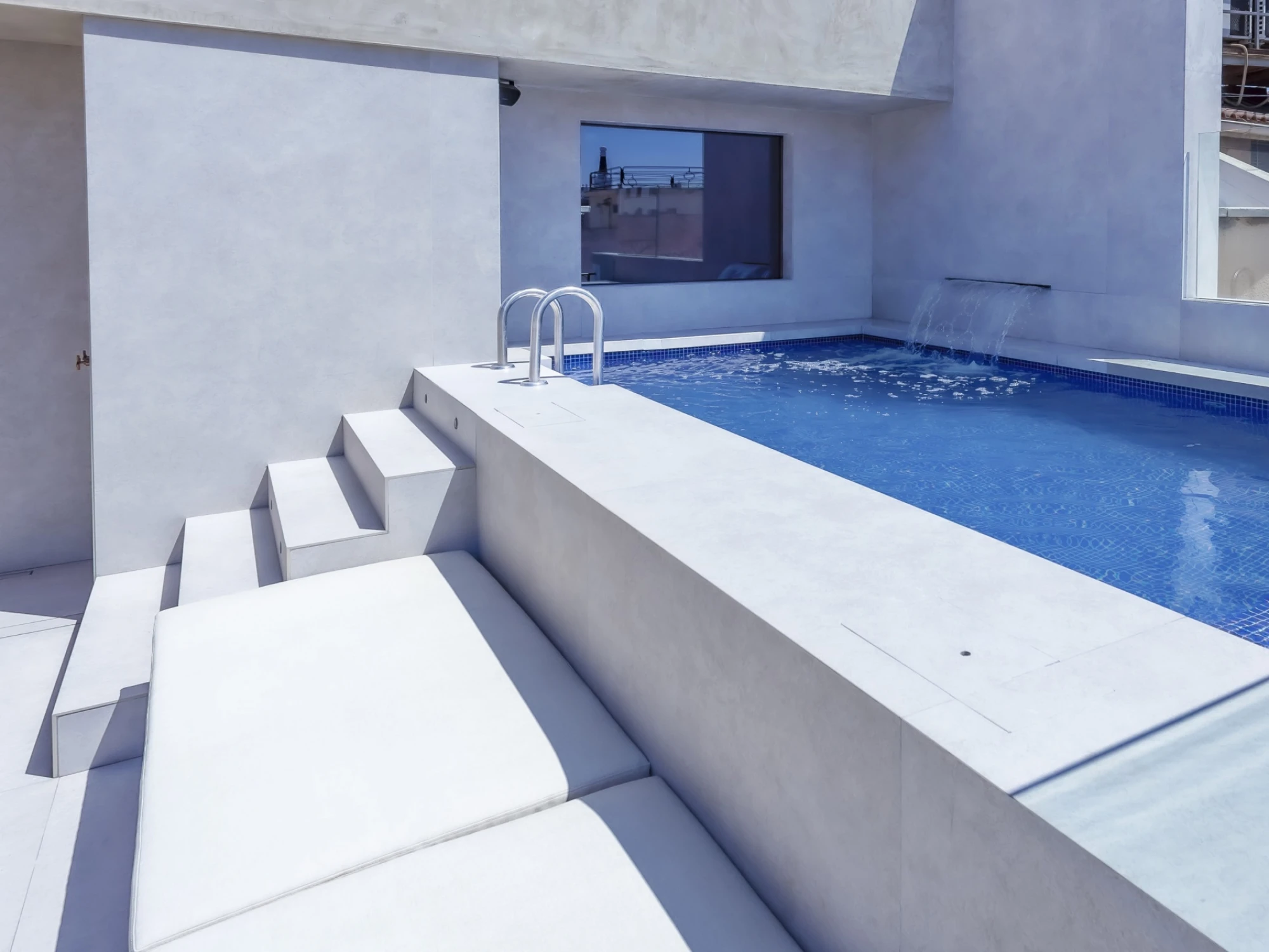 Se ve una piscina rectangular en la azotea con tumbonas blancas y una pared de hormigón alrededor.