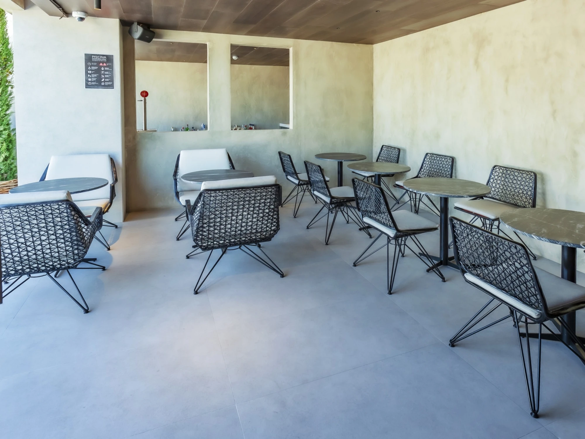 Se ve una terraza exterior con varias sillas y mesas metálicas, suelo de hormigón y paredes claras.