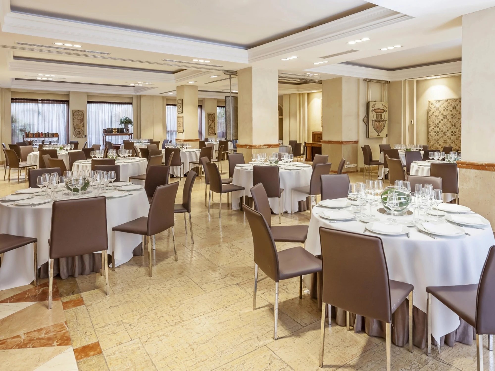 Se ve una sala de banquetes con mesas cubiertas y sillas alrededor, con iluminación de techo y columnas.