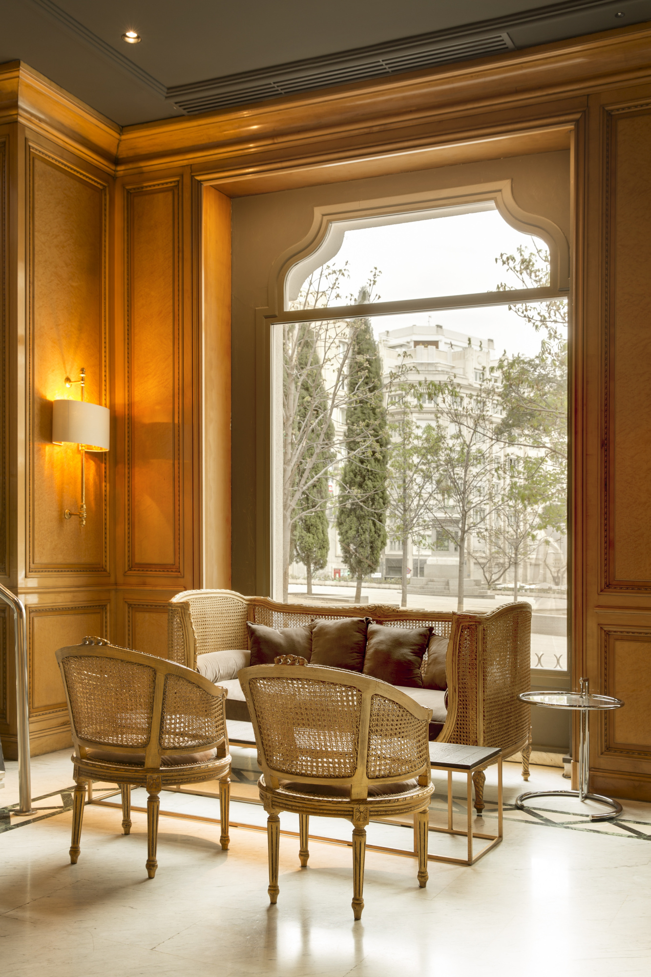 Se ve un área de lobby con sillones de mimbre, una mesa auxiliar y un gran ventanal con luz natural.