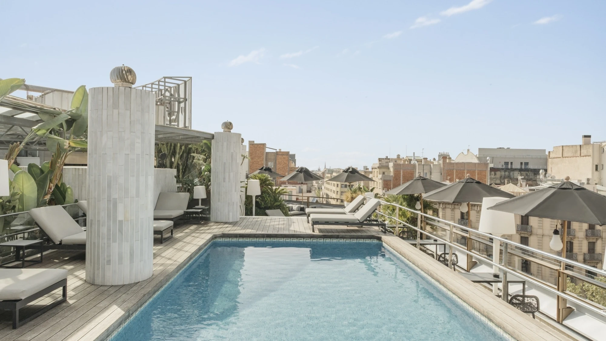 Piscina del Hotel Claris en Barcelona, vista desde la terraza con tumbonas elegantes y sombrillas, agua cristalina reflejando el cielo azul y vegetación que rodea el área creando un ambiente relajante y exclusivo.