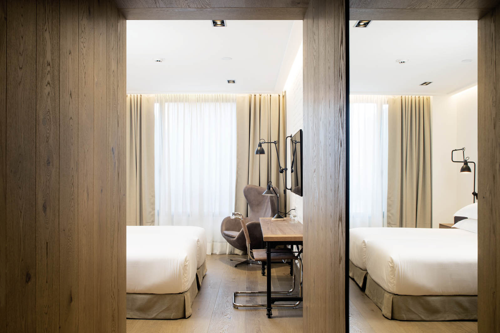 Se ven dos camas individuales con ropa de cama blanca, cortinas claras y una zona de trabajo con silla y mesa dentro de un dormitorio.