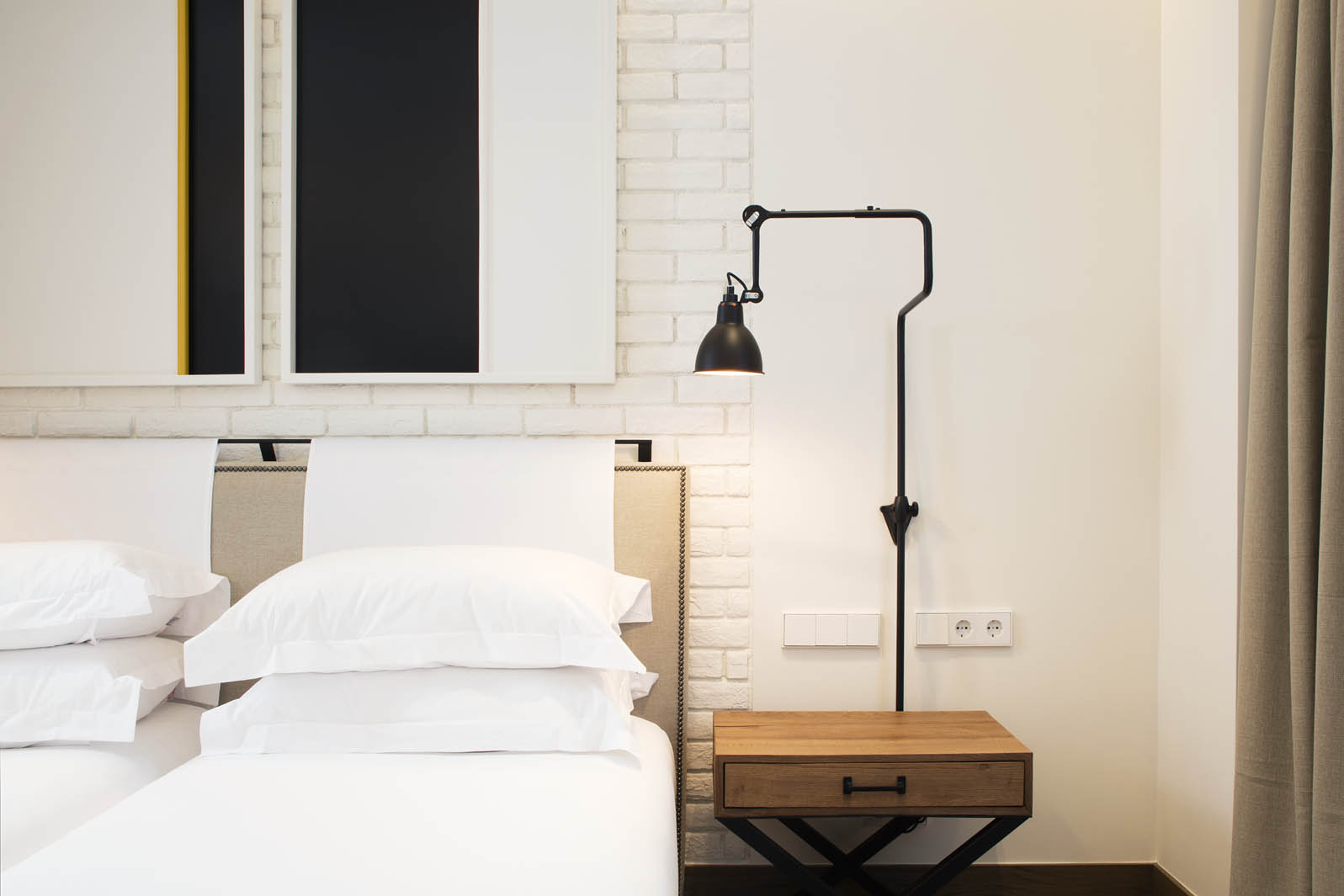 Se ve una cama con ropa de cama blanca junto a una mesita de madera y una lámpara de pie, con pared clara detrás.