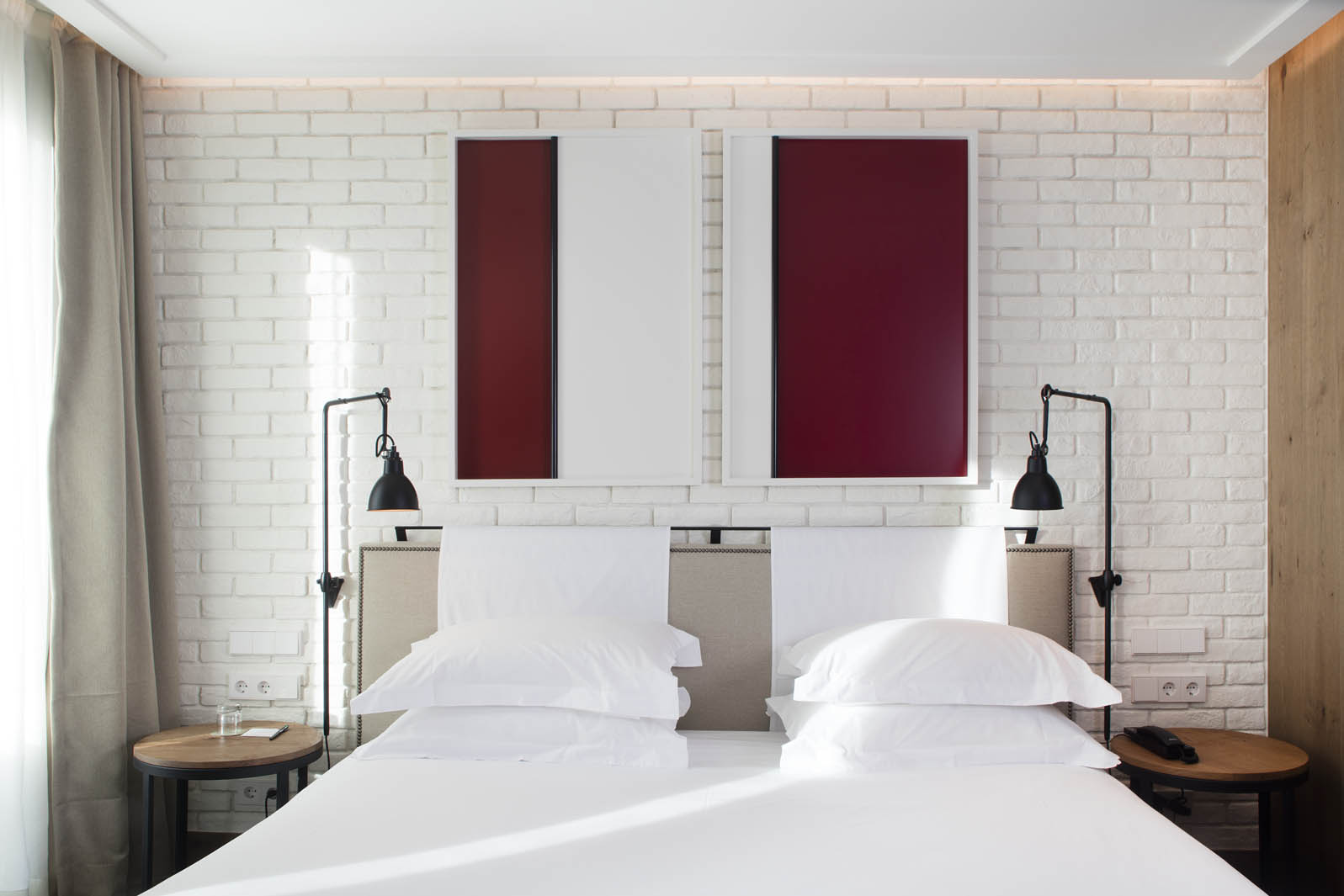 Cama doble con ropa de cama blanca, mesitas y lámparas sobre un fondo de pared de ladrillo blanco.