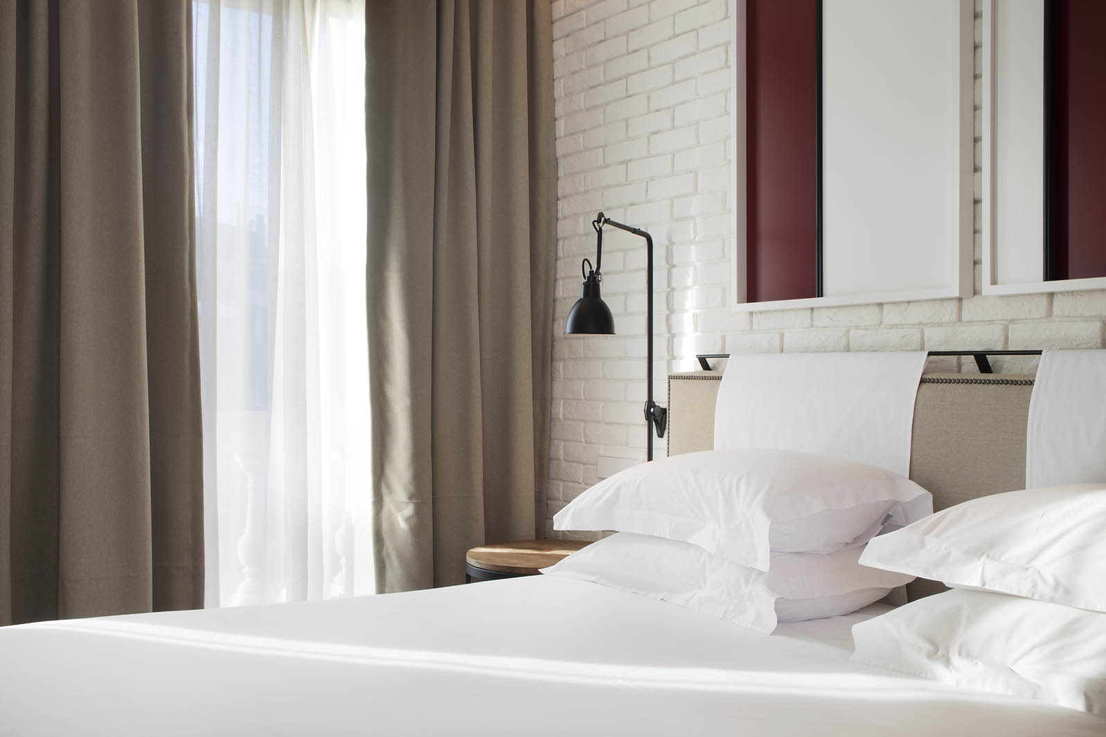 Se ve una cama con ropa de cama blanca y una cortina junto a una ventana, con un escritorio pequeño al fondo.
