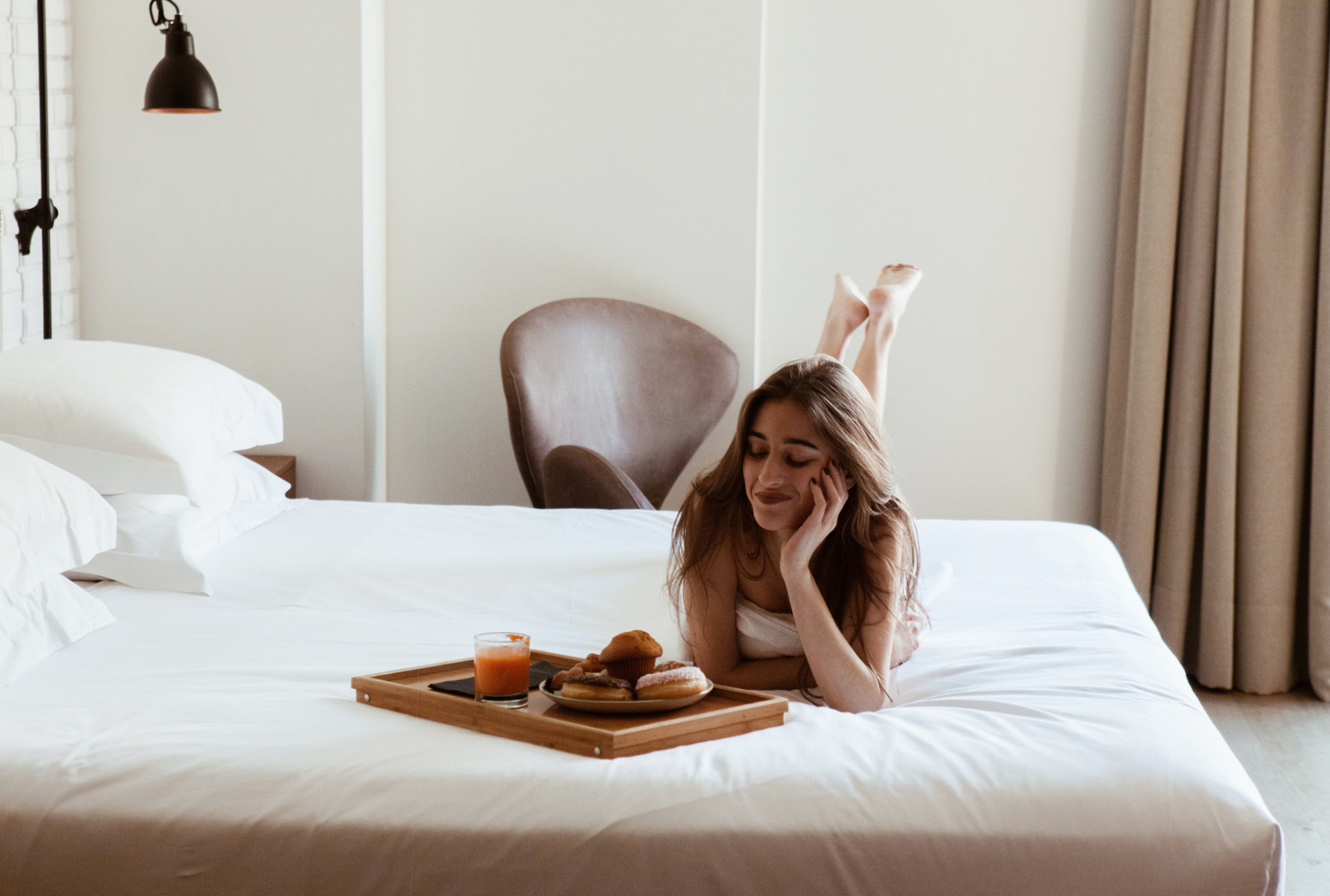 Una mujer desayuna en una cama con una bandeja sobre la sábana y una silla al fondo.